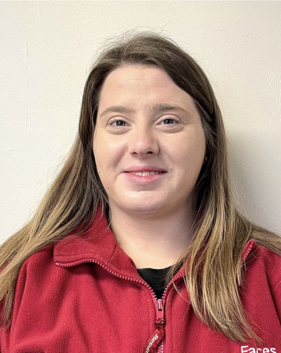 A brentwood preschool woman with long brown hair and fair skin wearing a red zip-up jacket standing against a light-colored wall.