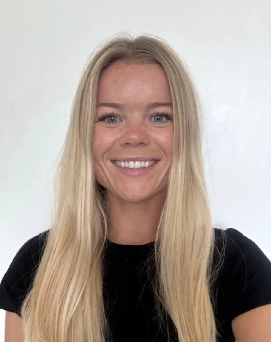 A smiling young office girl from a Brentwood preschool with long blonde hair, blue eyes, and light skin, wearing a black shirt, standing against a plain white background.