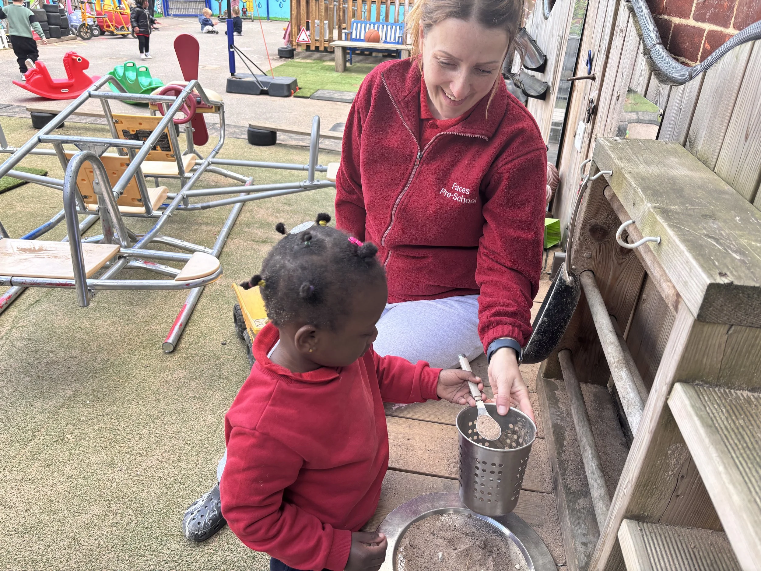 A young girl in a red hoodie with her hair in braids stands outdoors at a playground, pouring sand into a metal container held by a woman wearing a red jacket with 'Faces Pre-School' embroidered on it. Several children and playground equipment are visible in the background.