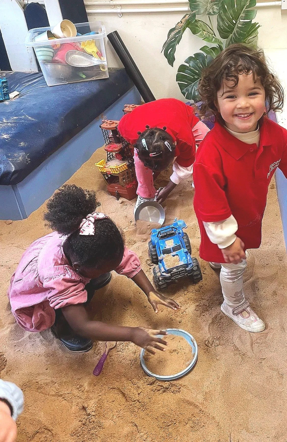 Three children playing in a sandbox with toys, a girl in a pink outfit, a girl in a red shirt with curly hair, and a child in a red hoodie with a caterpillar design. One is smiling at the camera, and others are engaged with toys.