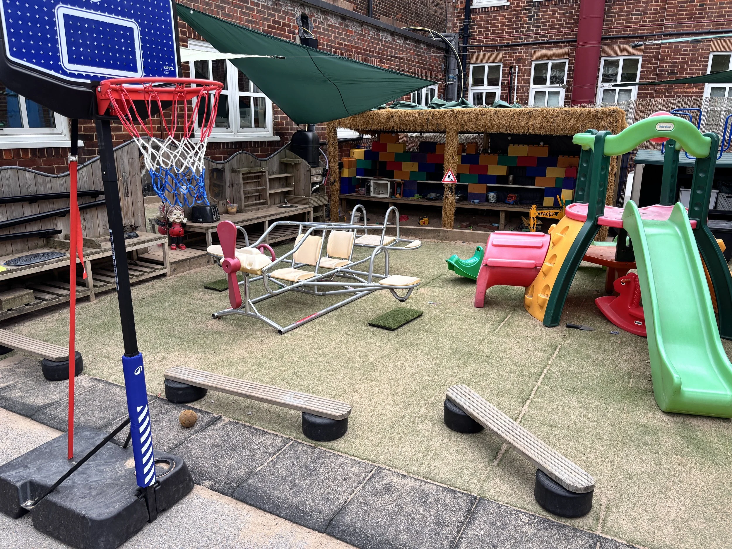 Children's outdoor play area with a small basketball hoop, green slide, and a shaded wooden playhouse with colorful brick wall design.