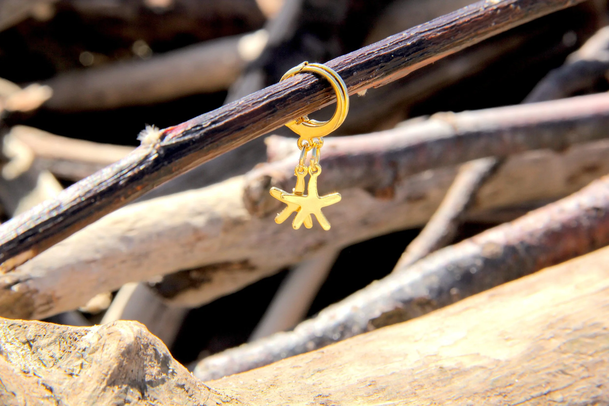 A pair of 18K gold-plated brass starfish charm huggie hoop earrings suspended from a driftwood stick on the beach.