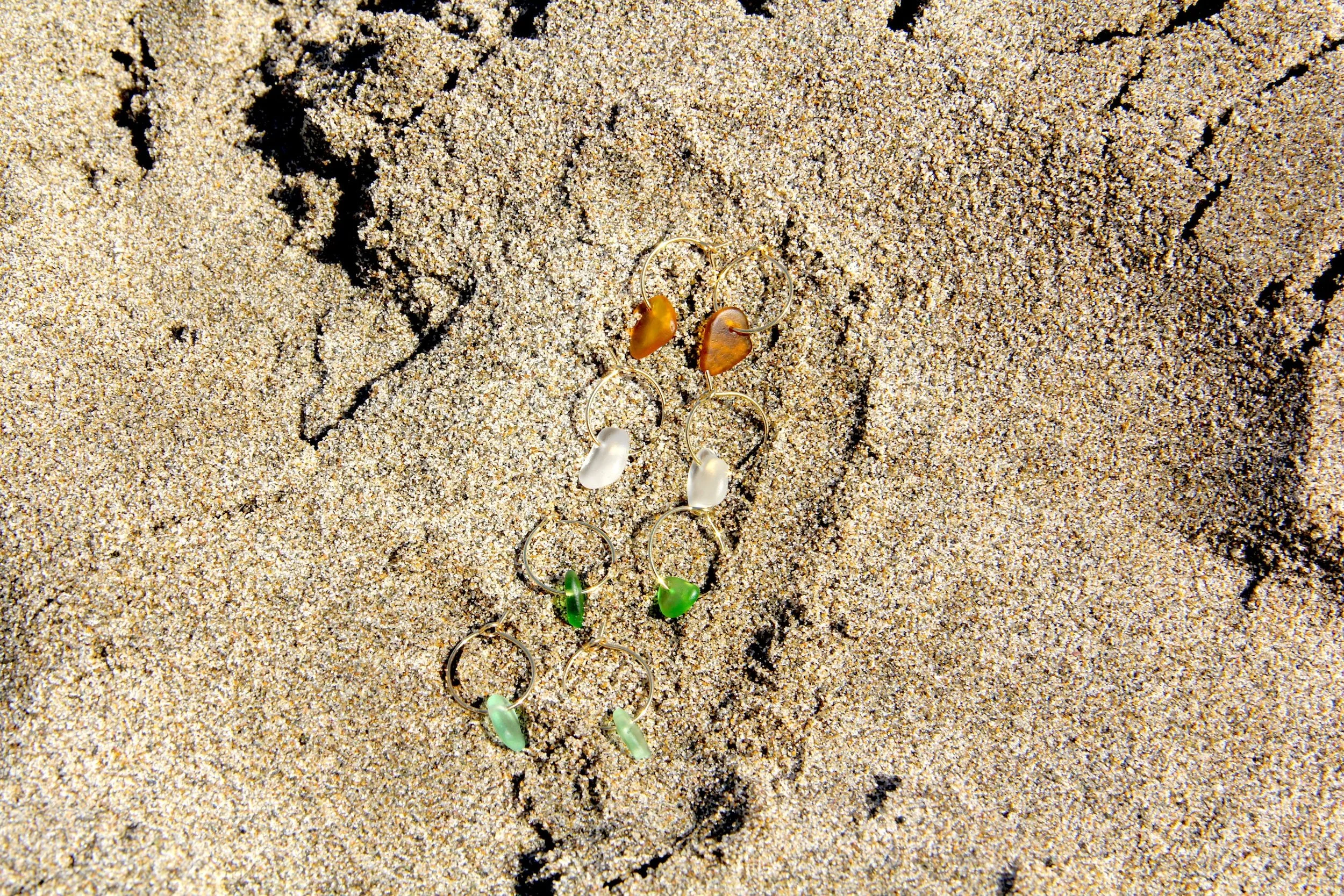 Four pairs of gold stainless steel sea glass hoop earrings lying in beach sand.