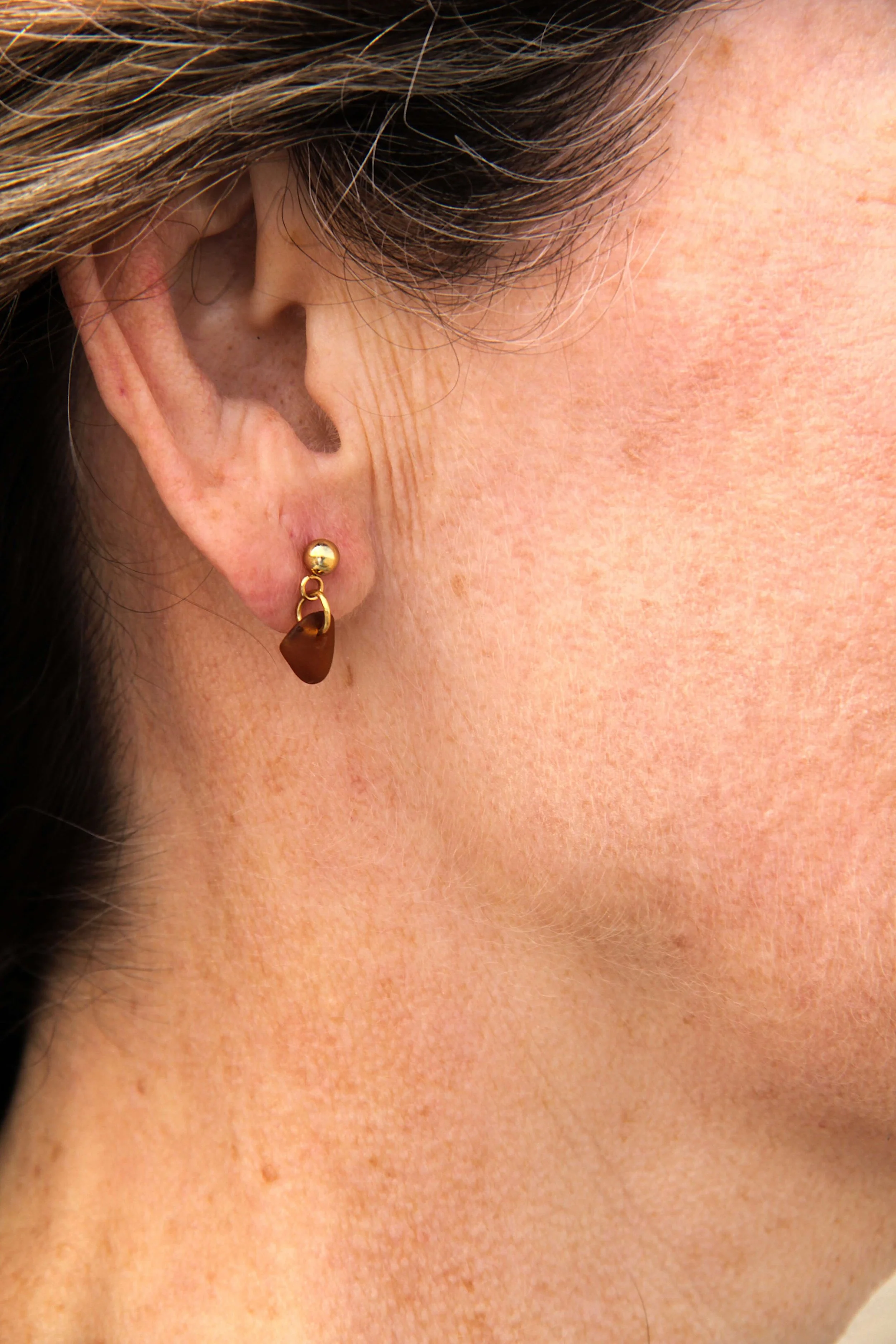 Close up of woman wearing a 18K gold-plated stainless steel brown sea glass stud earring on her ear.
