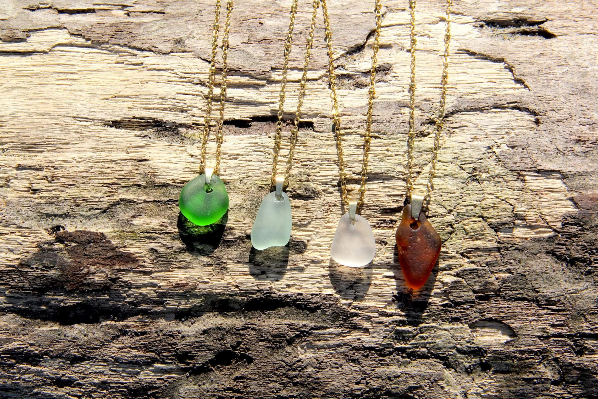Four genuine ocean-tumbled sea glass pendant necklaces laying on a driftwood log on the beach.