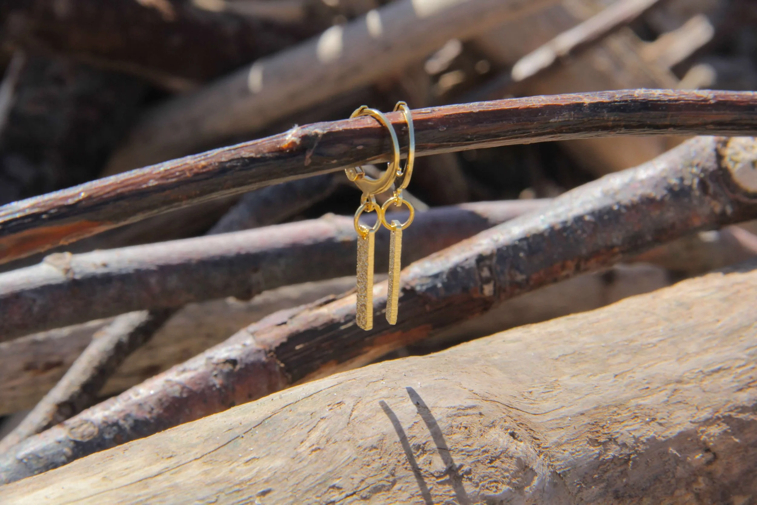 A pair of 18K gold-plated brass textured bar charm huggie hoop earrings suspended from a driftwood stick on the beach.