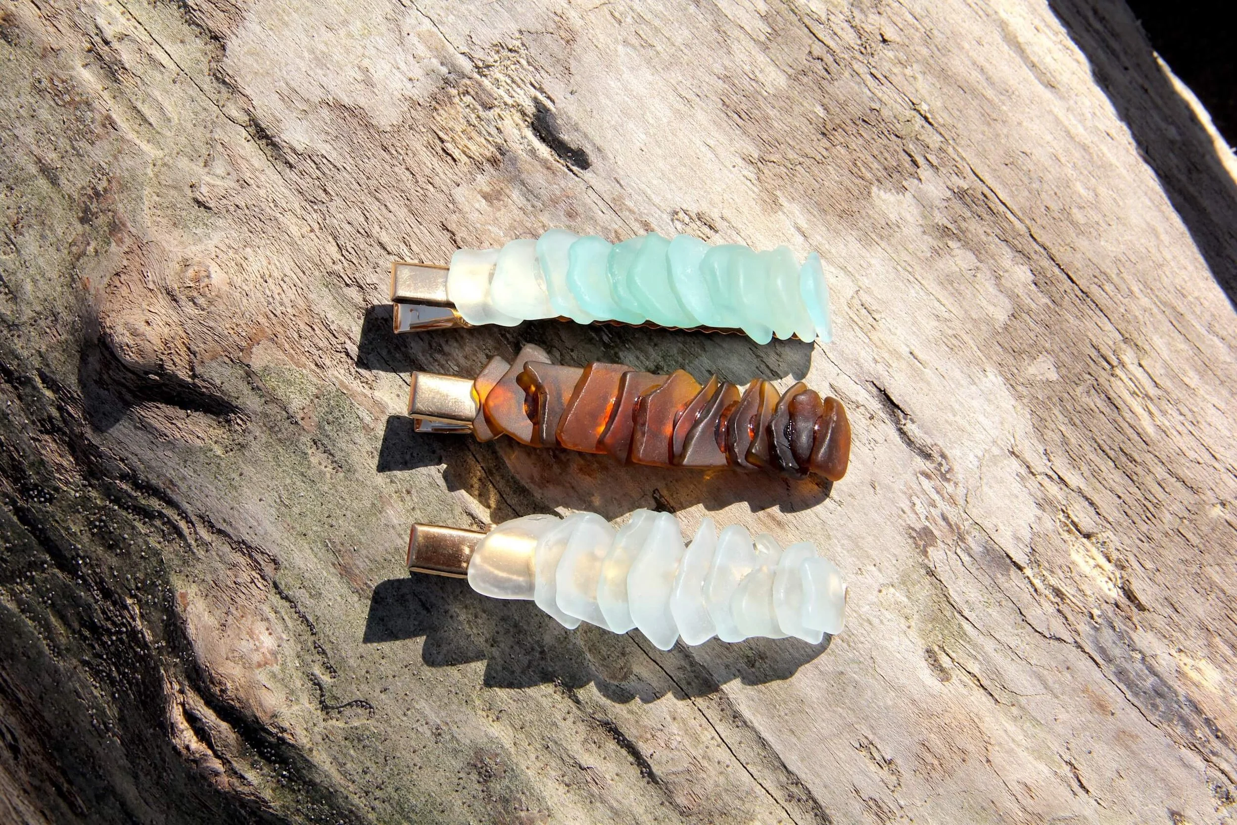 Three genuine ocean-tumbled sea glass gold hair clips laying on a driftwood log on the beach.