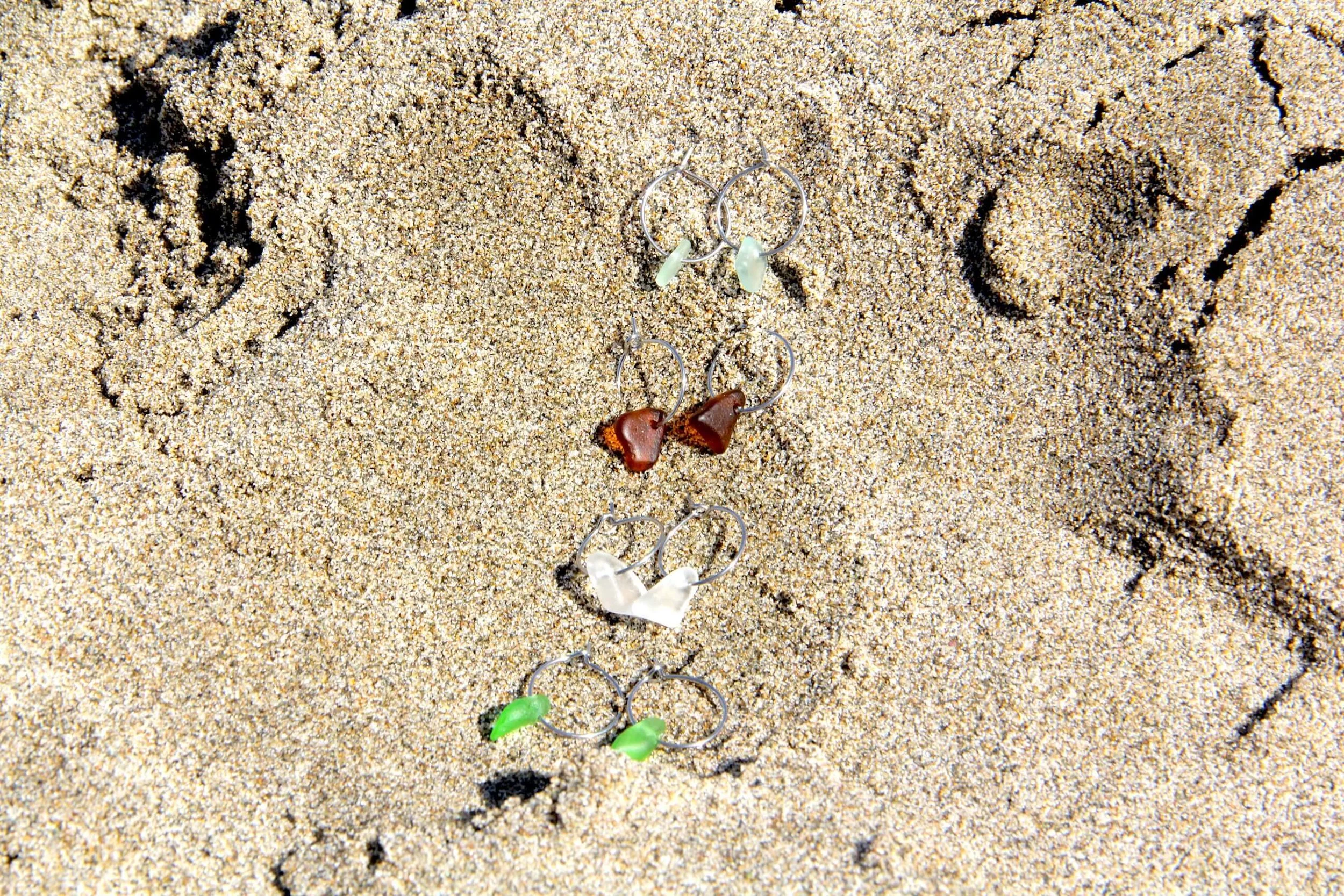 Four pairs of silver stainless steel sea glass hoop earrings lying in beach sand.