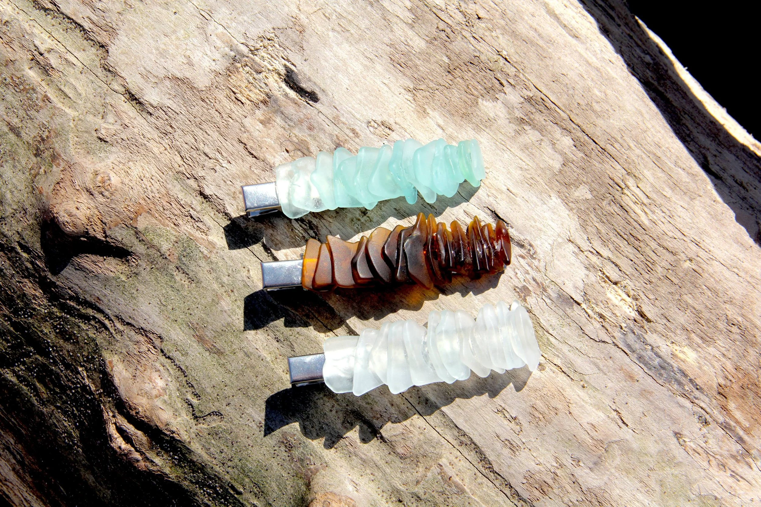 Three genuine ocean-tumbled sea glass silver hair clips laying on a driftwood log on the beach.