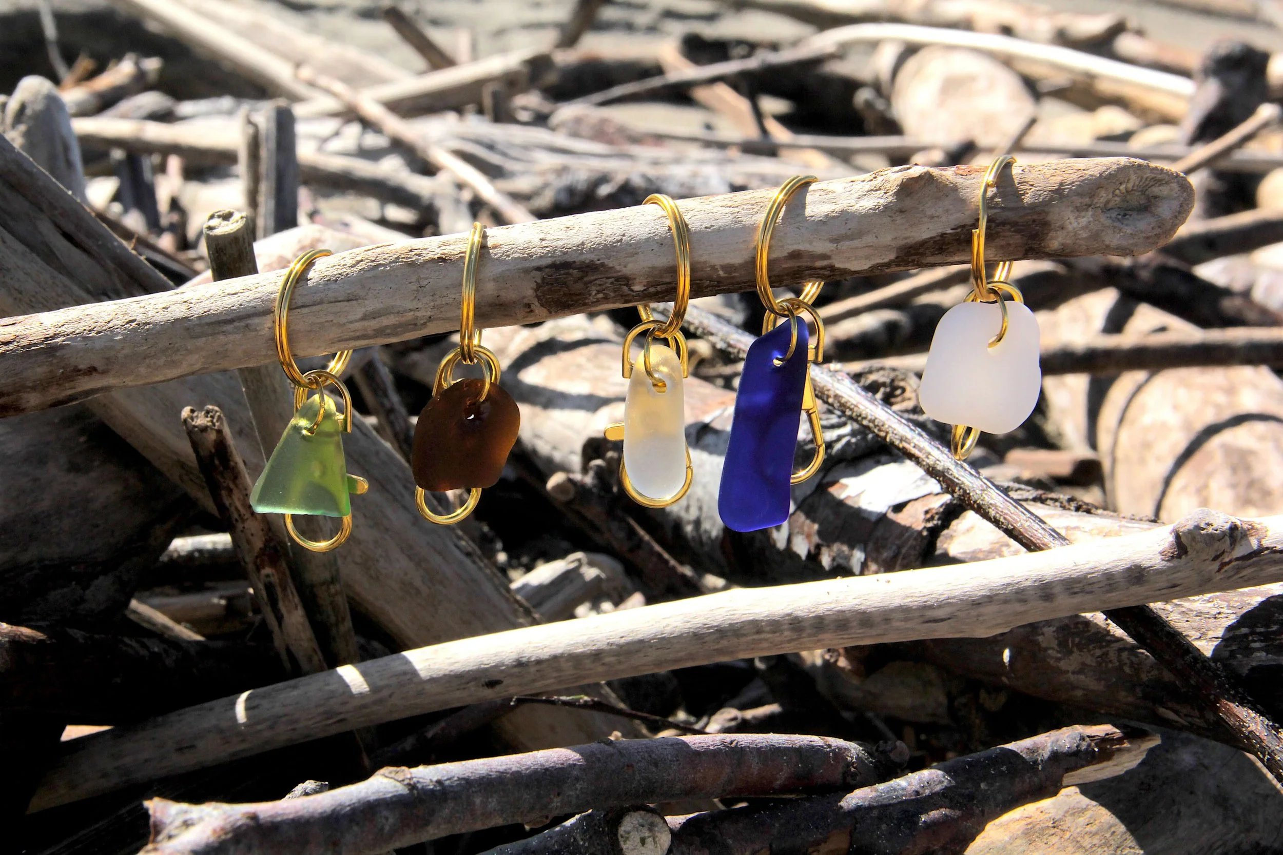 Green, brown, clear, blue, and white genuine ocean-tumbled sea glass keychains suspended from a driftwood stick on the beach.