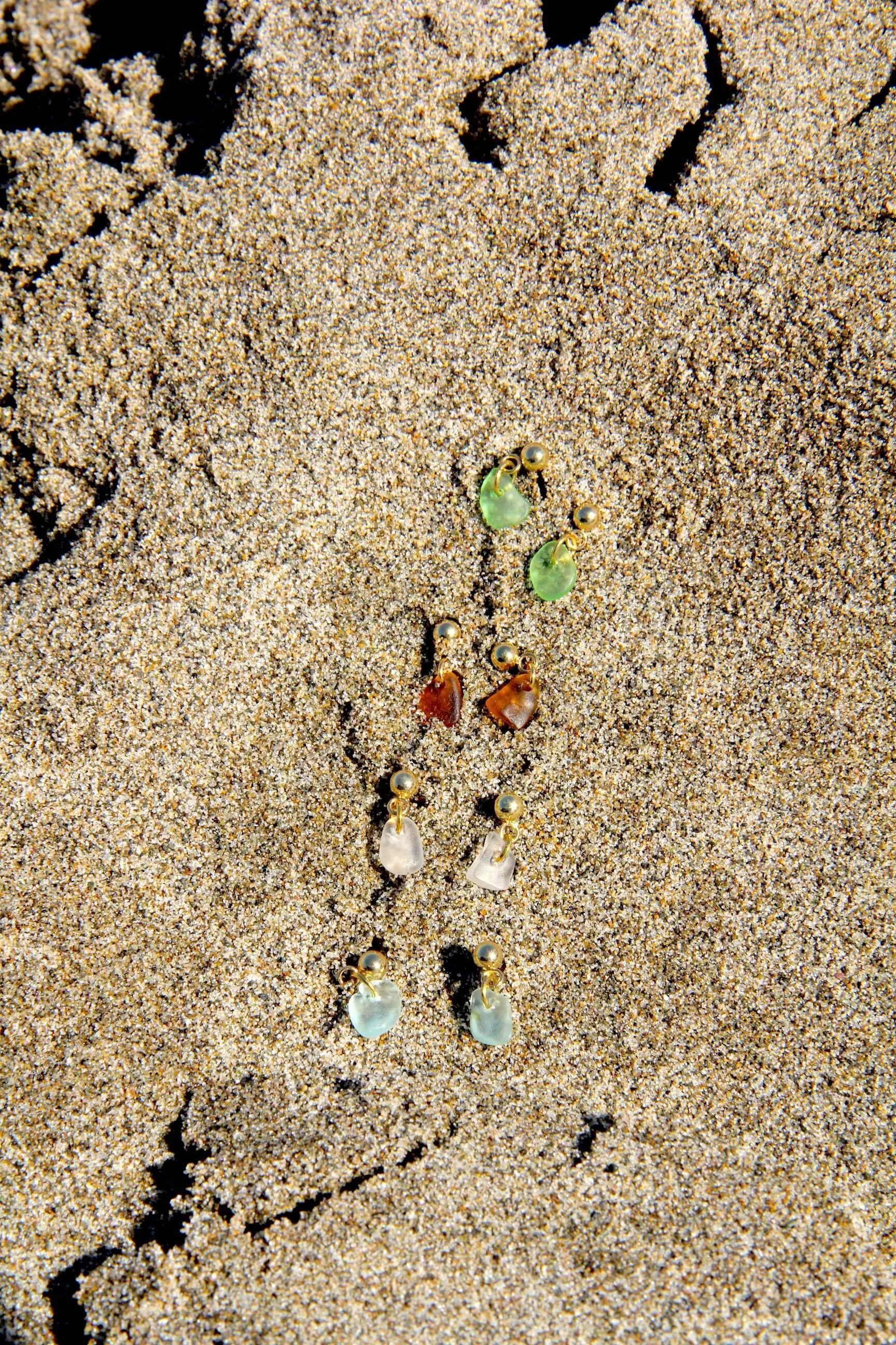 Four pairs of 18K gold-plated stainless steel sea glass stud earrings lying in beach sand.