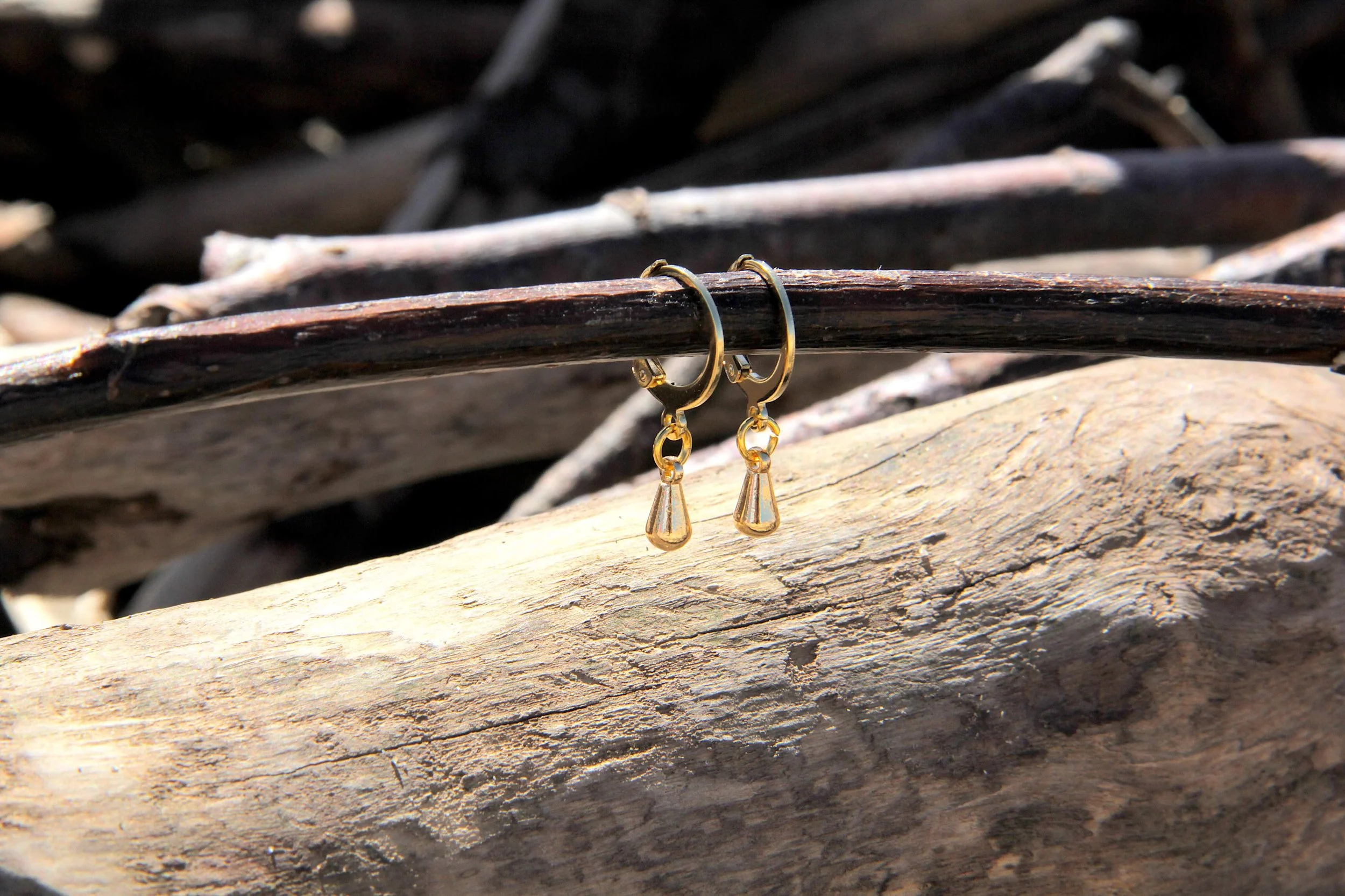 A pair of 18K gold-plated brass water droplet charm huggie hoop earrings suspended from a driftwood stick on the beach.
