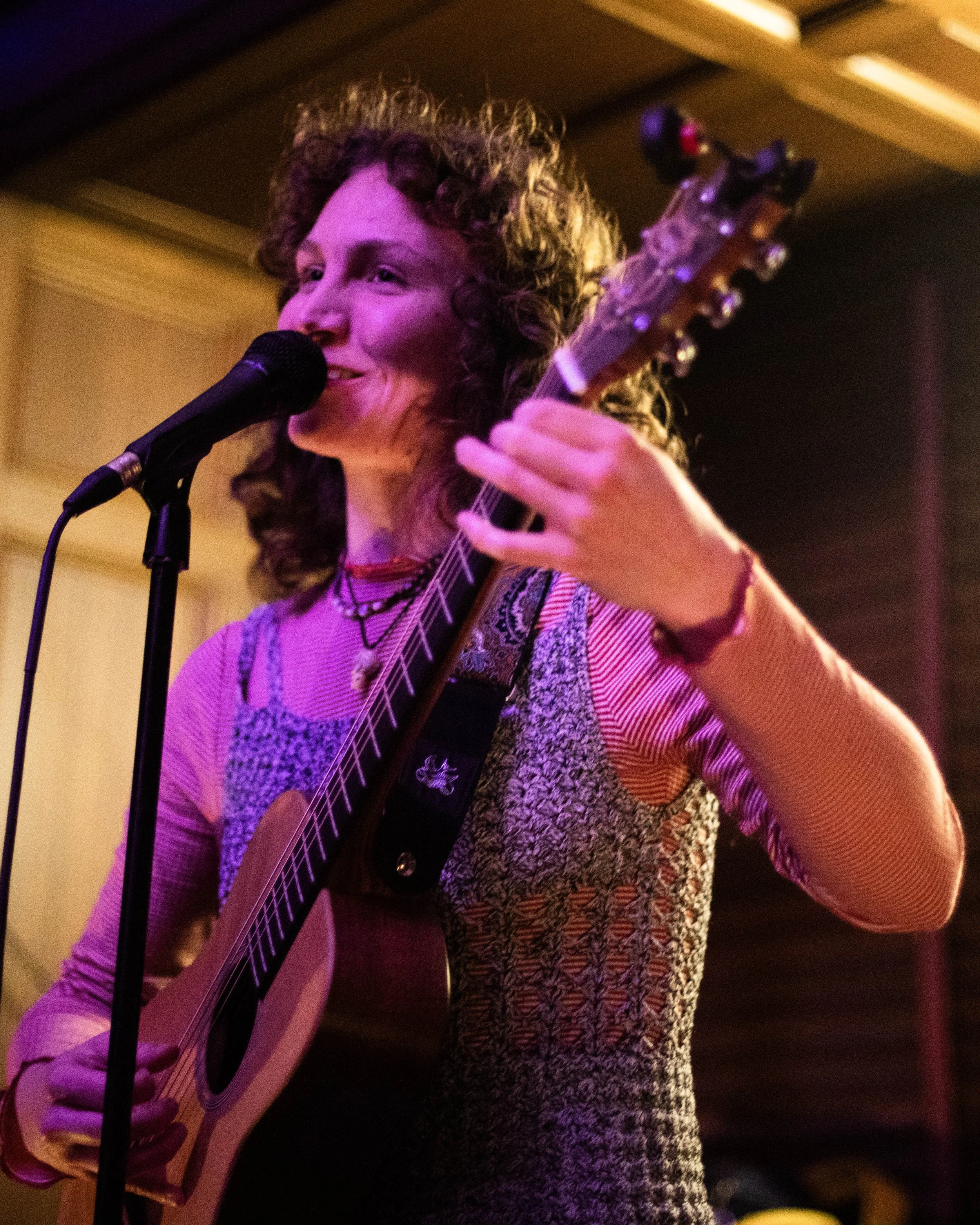 A woman with curly hair singing into a microphone while playing an acoustic guitar, wearing a patterned blouse, in a dimly lit room.