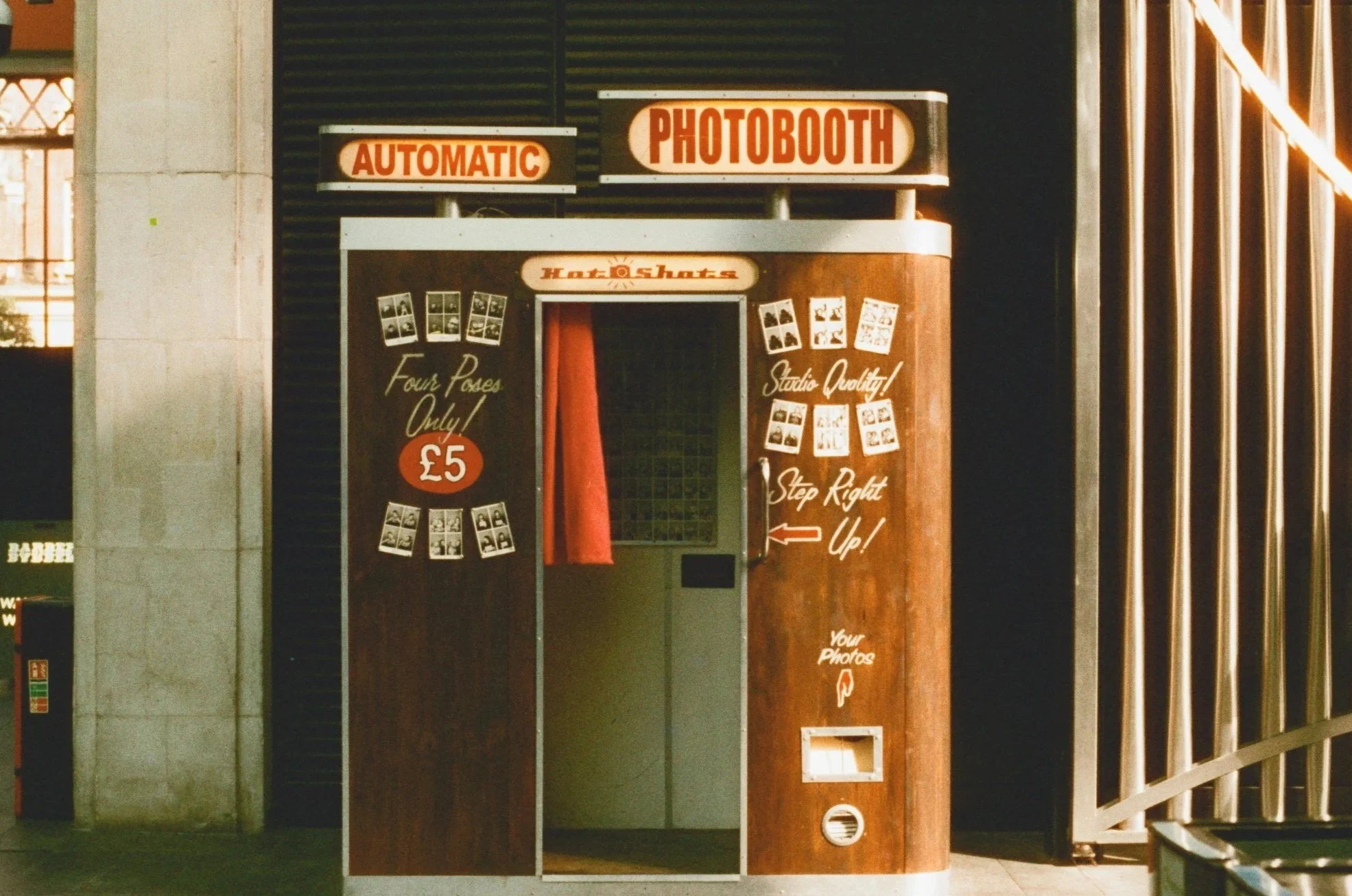 An outdoor photo booth with signs reading 'Automatic Photobooth', 'Hot Shots', and instructions to step right up for photos costing £5. The booth has four photo strips on display and a red curtain inside for photo taking.