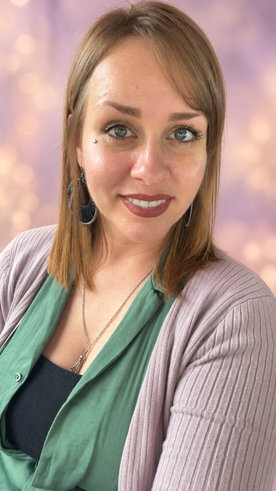 Close-up portrait of a woman with shoulder-length reddish-brown hair, blue eyes, wearing earrings, a necklace, a green blouse, and a beige cardigan, smiling softly against a pink and purple bokeh background.