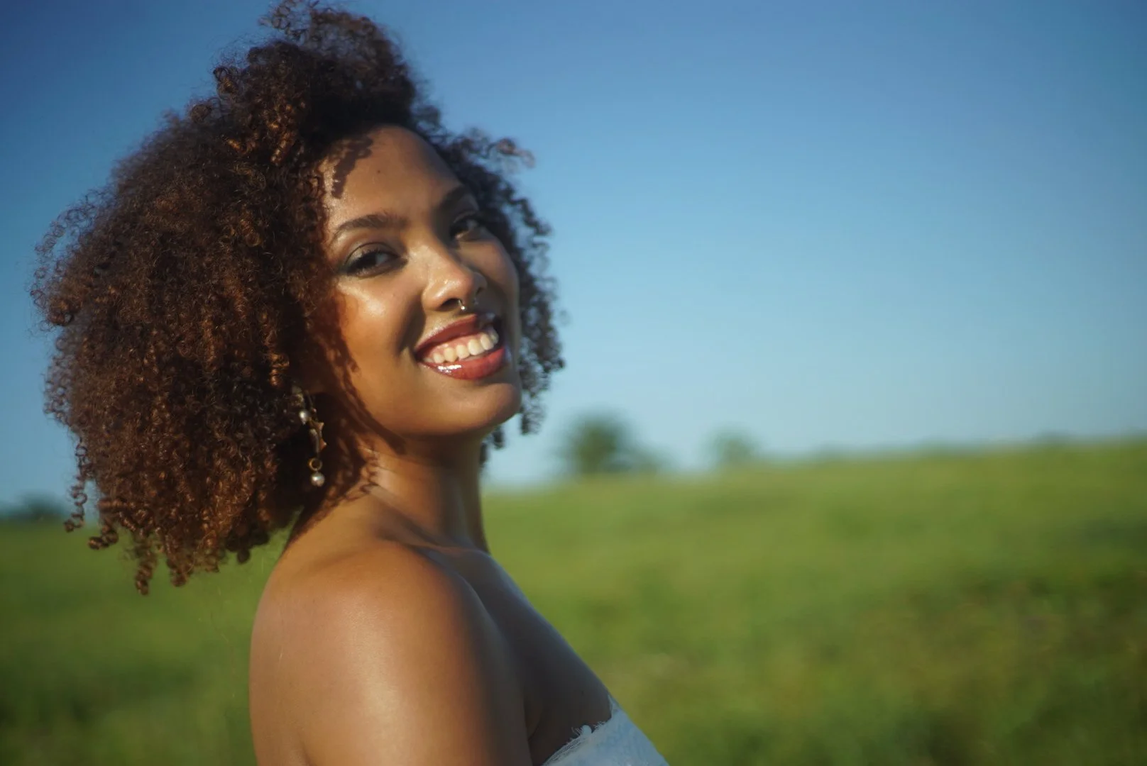 A woman with curly hair and a nose ring smiling in an outdoor field under a clear blue sky.