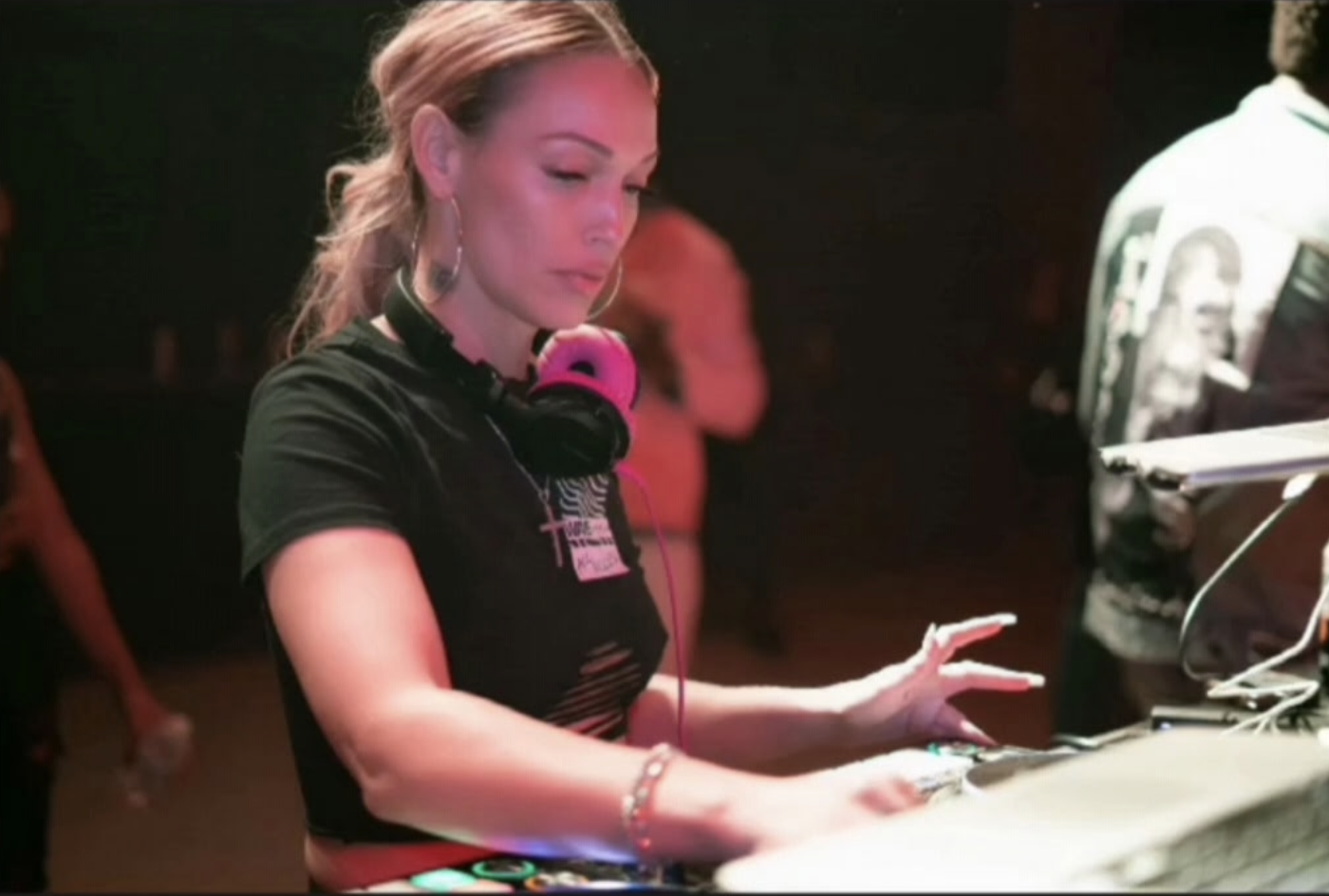 A woman DJing at a dance party with headphones around her neck, wearing a black shirt and silver earrings, focused on her equipment.