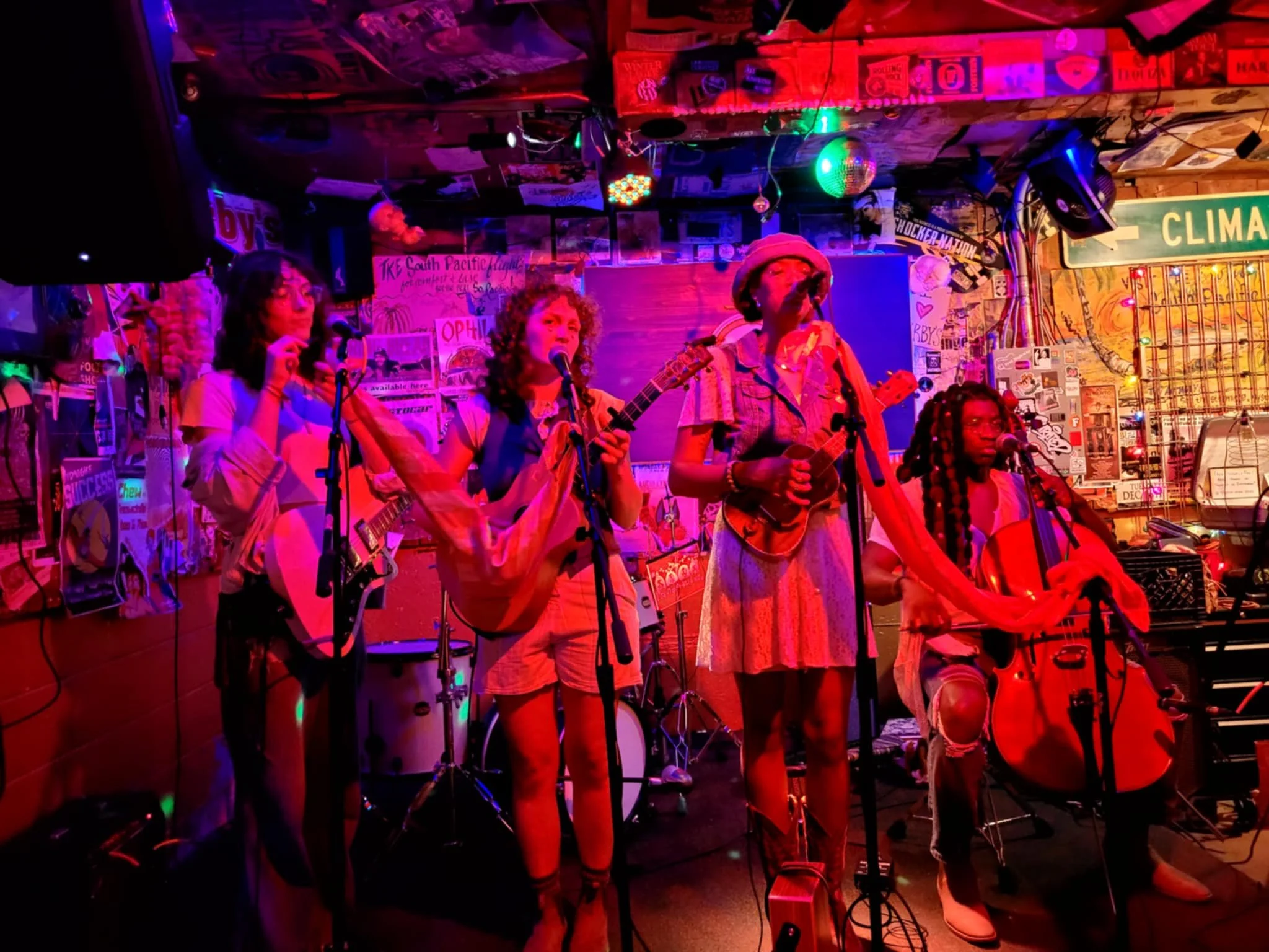 Four women performing on a small stage with musical instruments and microphones, under colorful stage lighting in a cozy, decorated venue.