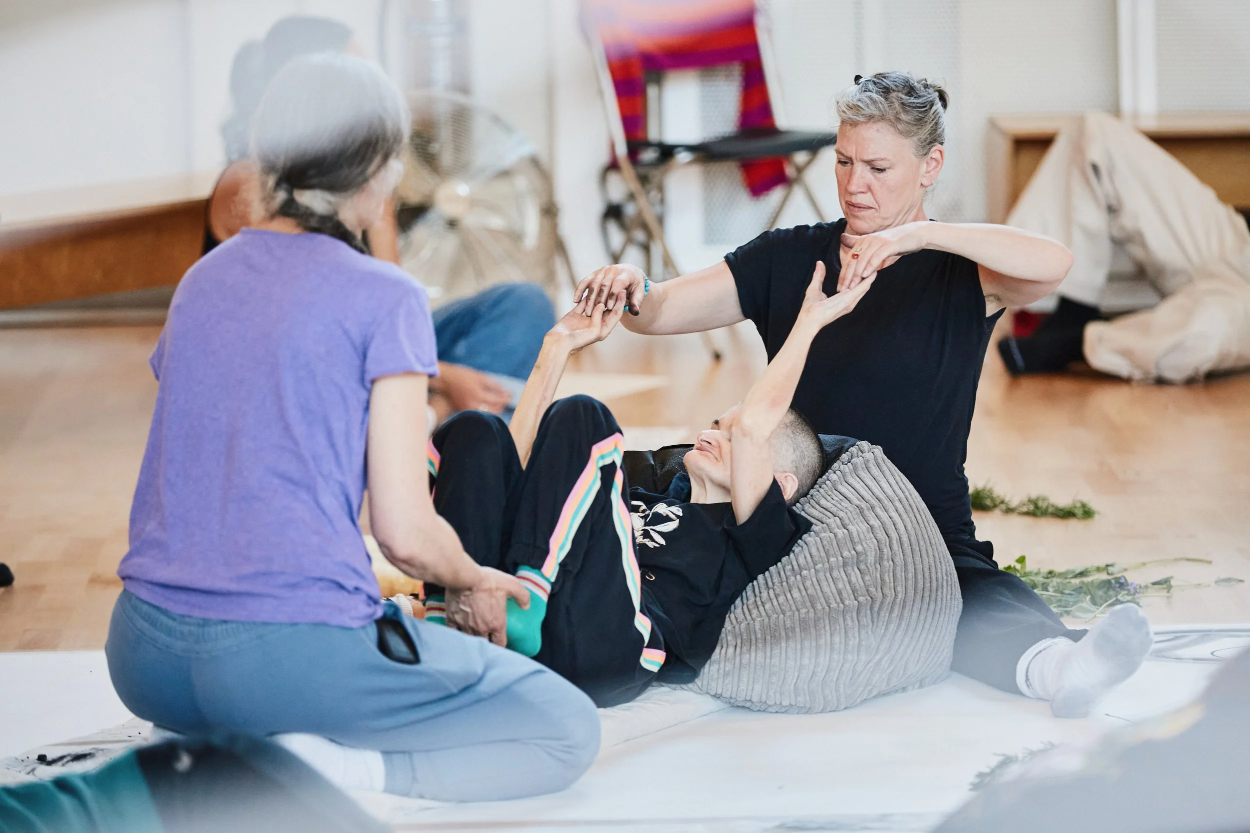 A woman lies between two other women, one woman holds her feet, the other woman holds her hands. They are dancing.