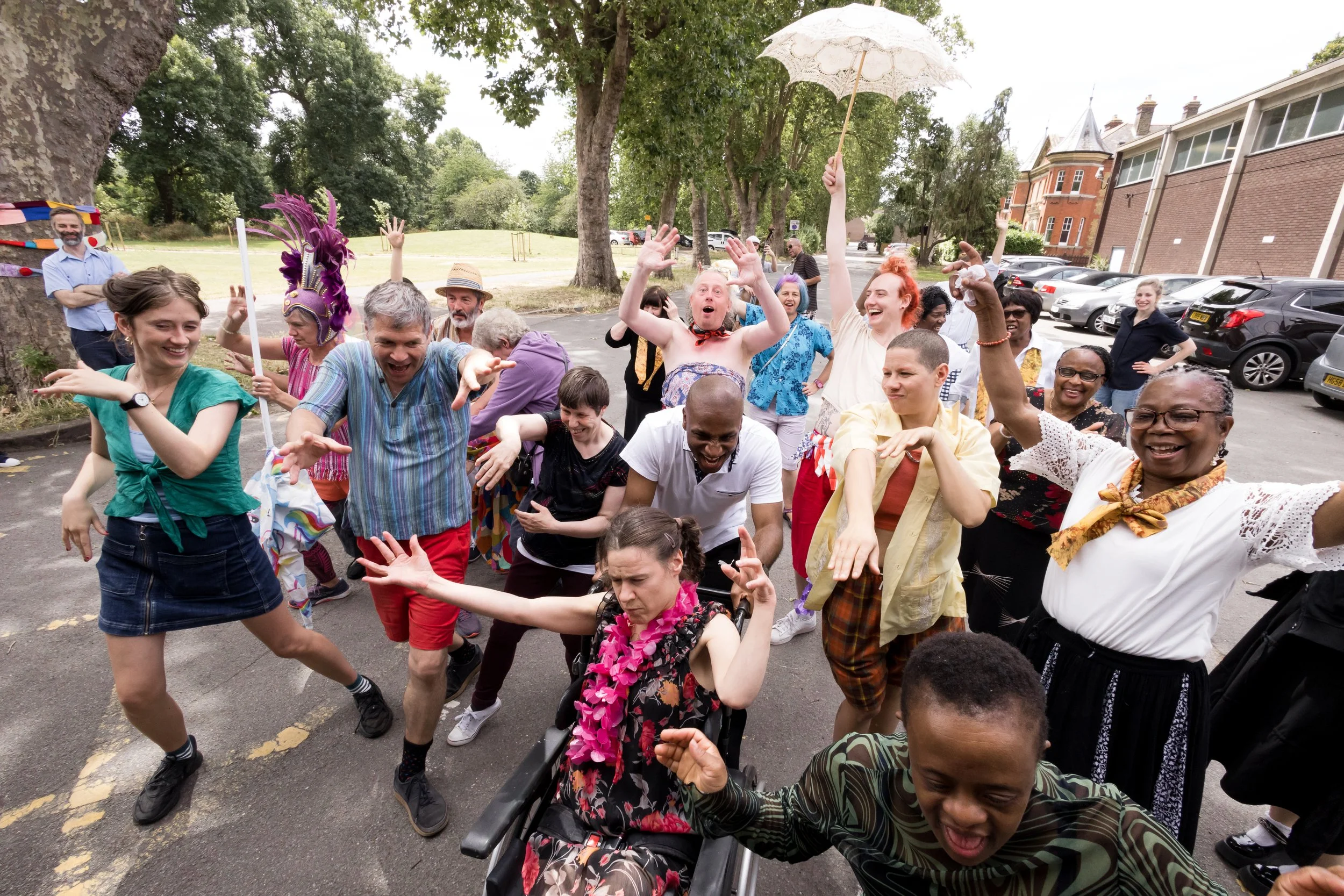 A group of around 20 people parading down a street next to a park with their arms in the air, smiling and dancing.