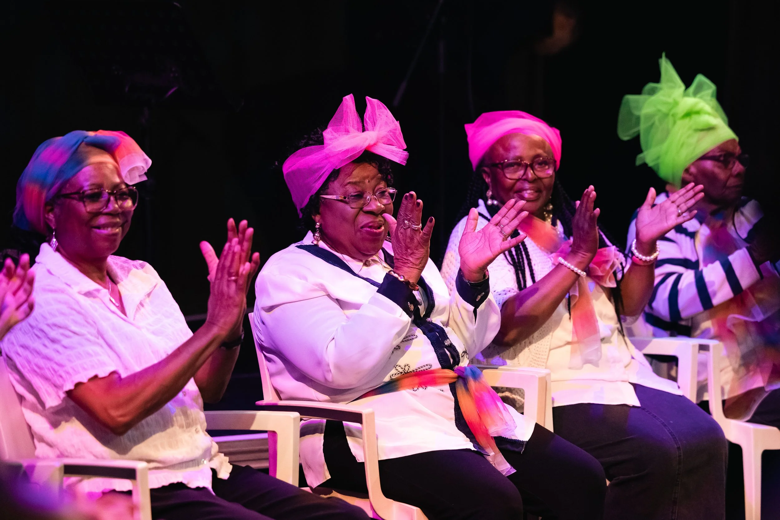 A row of 4 older women who are sitting, smiling and clapping their hands. All are wearing head scarfs and lit by a pink light on stage.