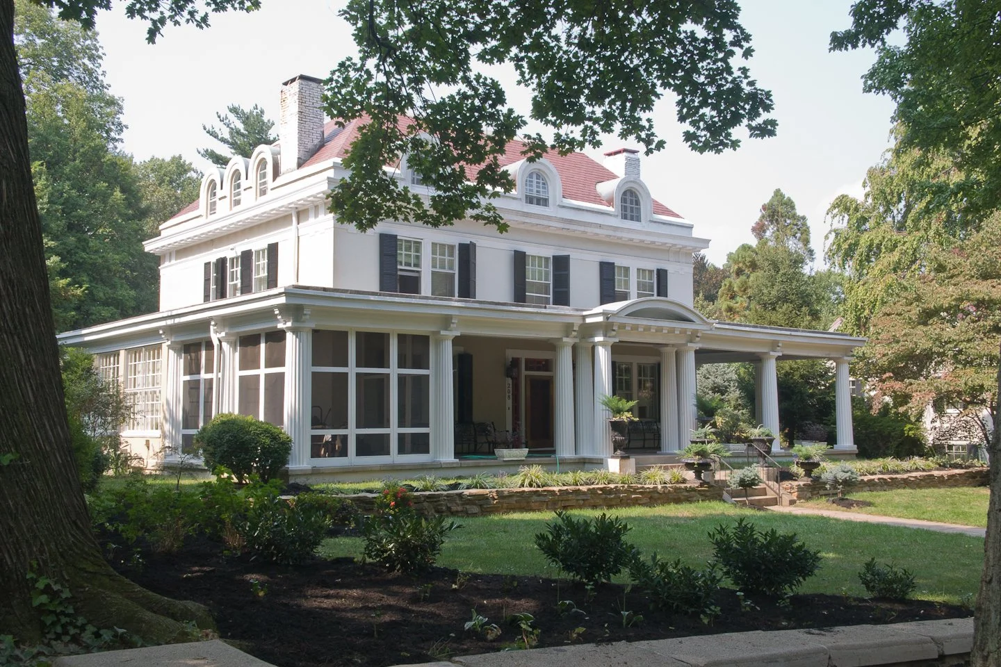 A large, white, three-story house with black shutters, a red roof, and a wraparound porch with columns, surrounded by trees and a landscaped yard.