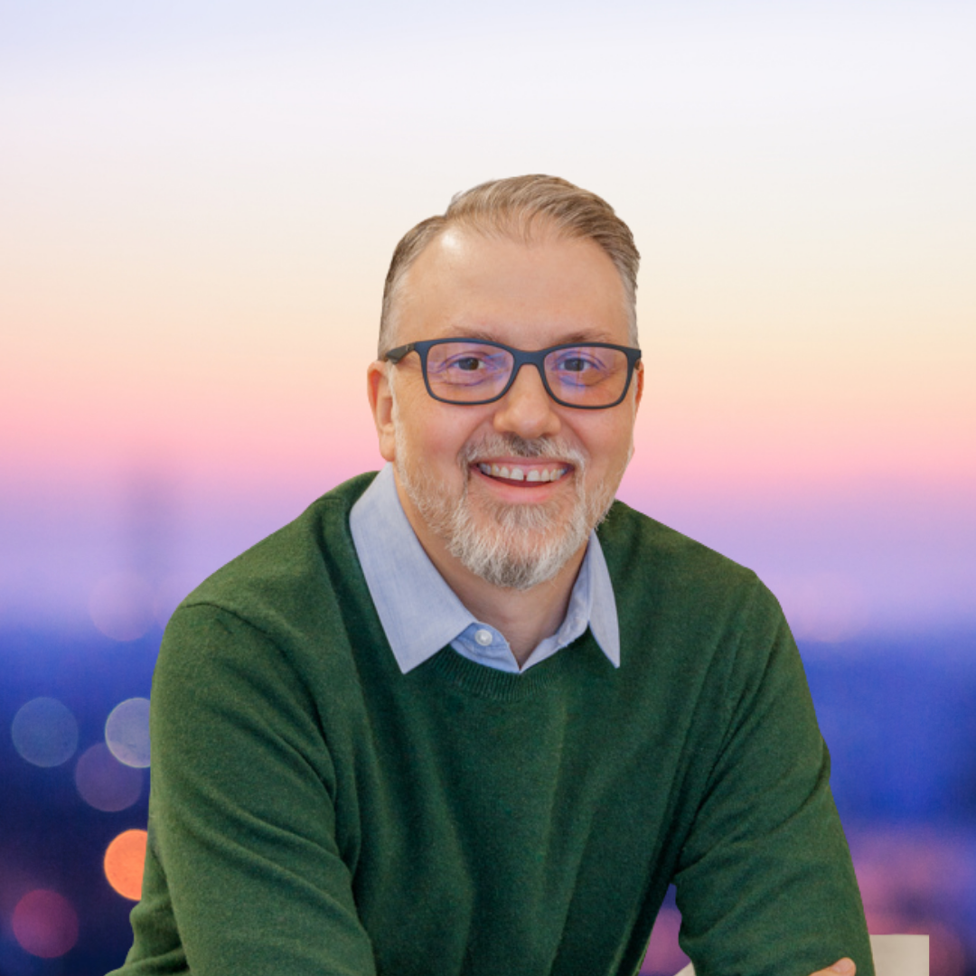 A smiling middle-aged man with short, graying hair and glasses, wearing a green sweater and light blue collared shirt, against a blurred pastel sunset background.