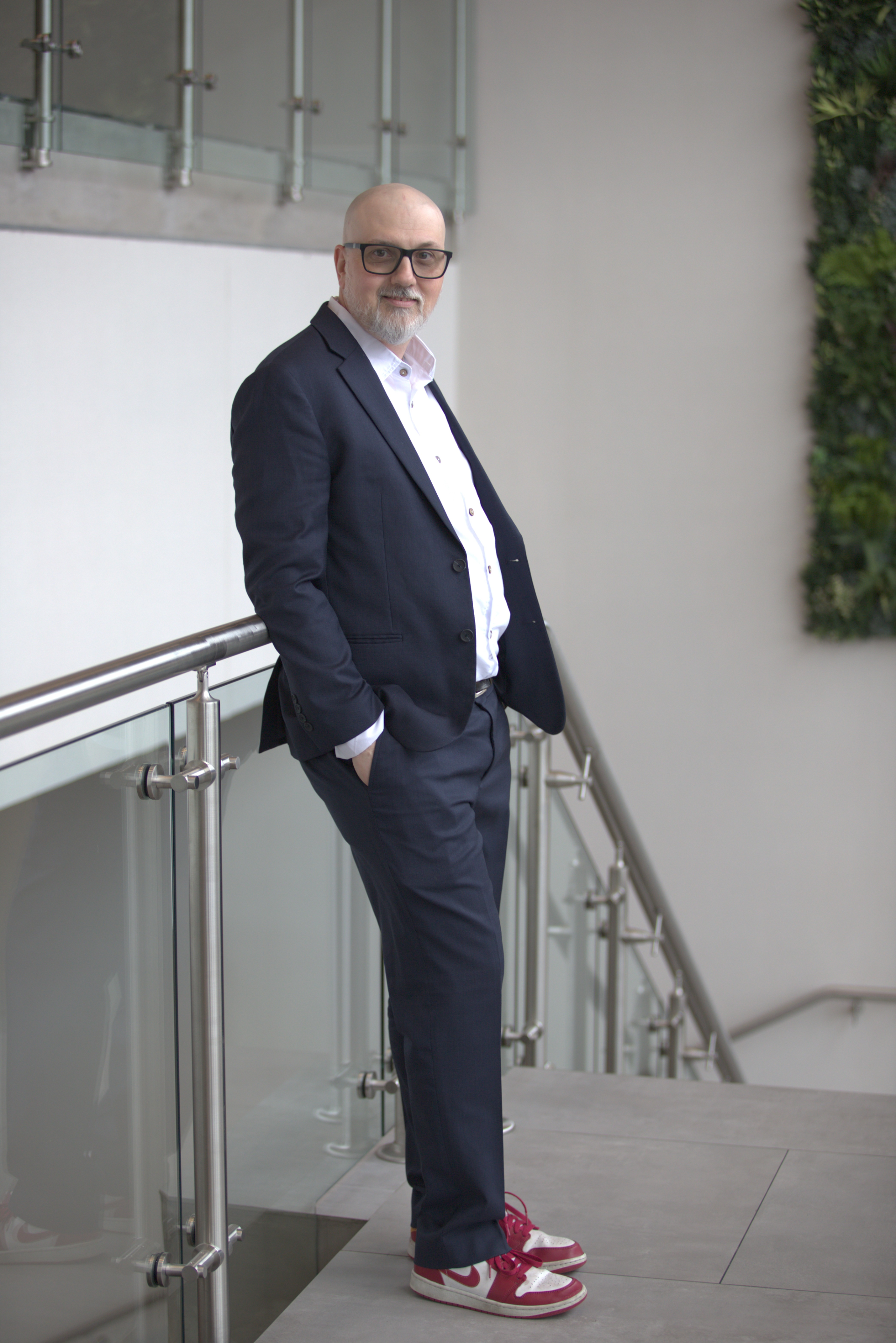 A man with glasses, a white beard, dressed in a navy suit, white shirt, and colorful sneakers, stands by a railing in an indoor setting with a white wall and greenery.