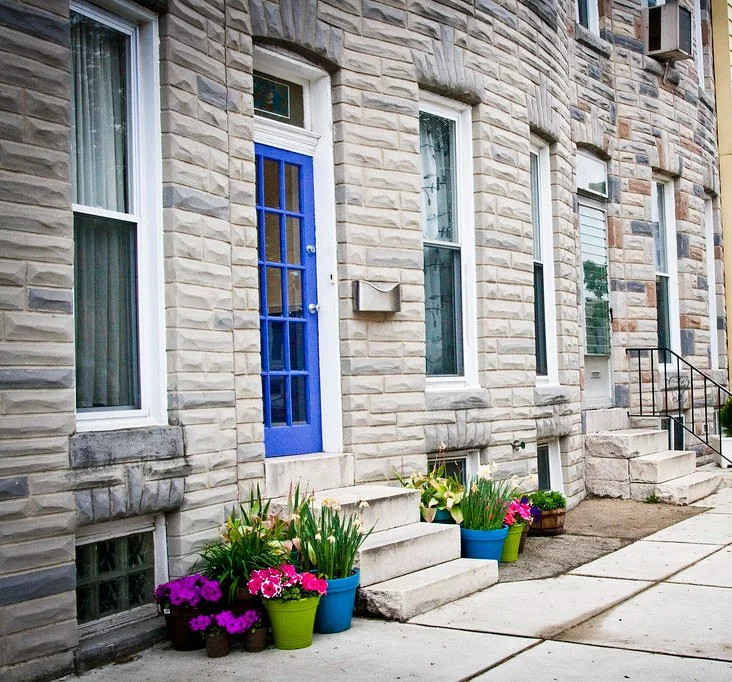 Stone apartment building facade with blue front door, multiple windows, potted plants and flowers along the sidewalk.