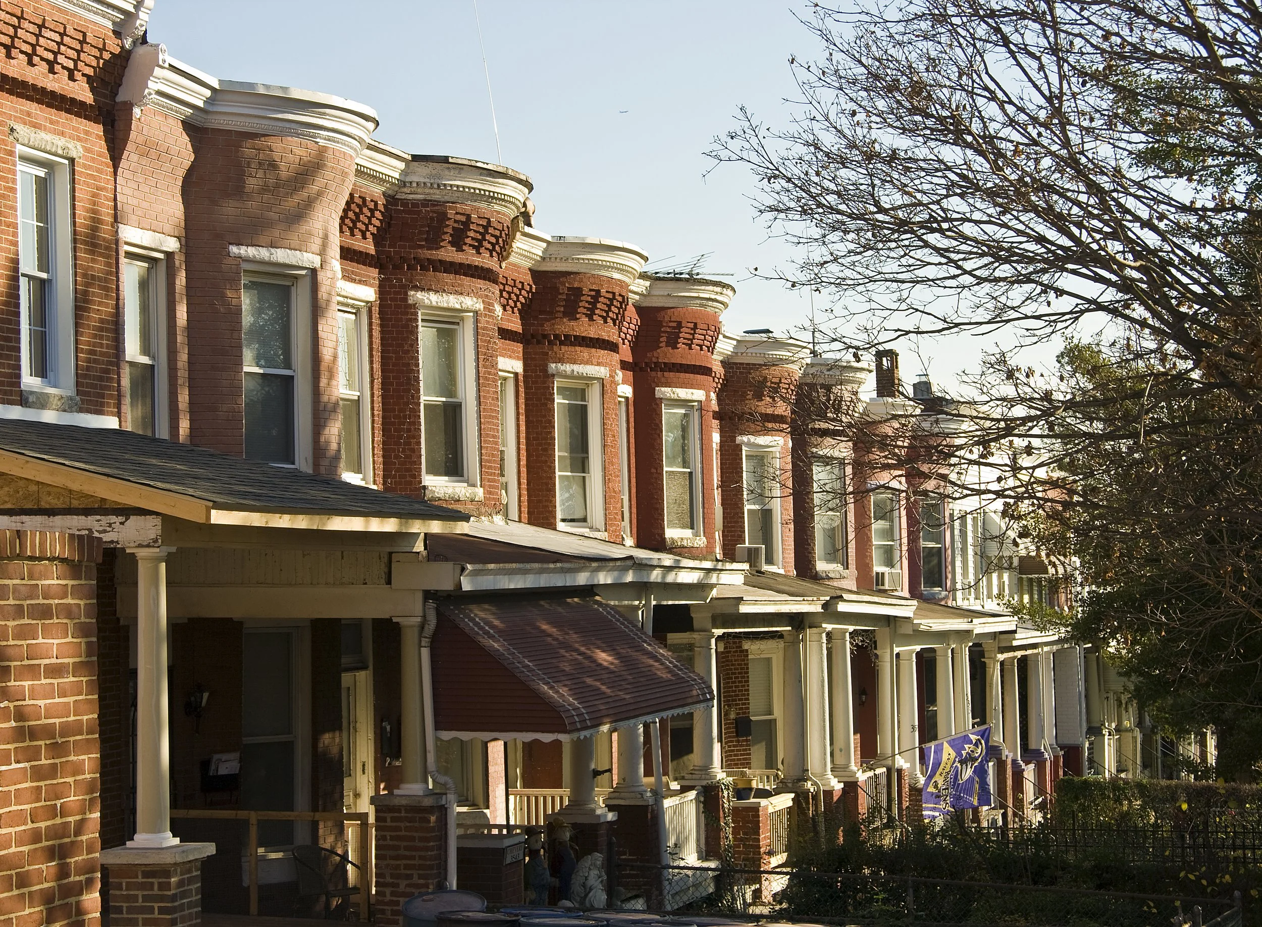Row of historic brick row houses with large front porches, white trim, and bay windows, decorated with flags, in an urban neighborhood during daylight with a clear sky and trees.