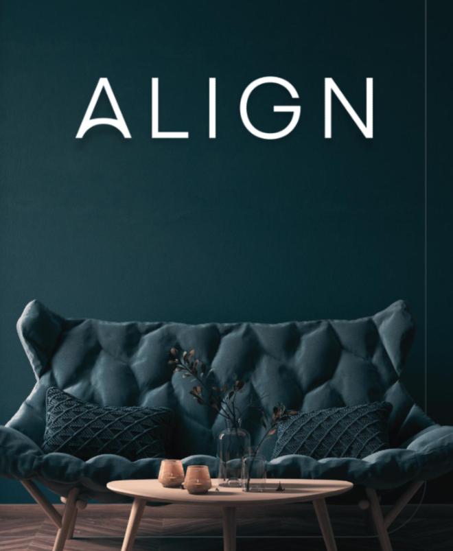 A living room with a dark teal wall displaying the word "ALIGN" in large white letters. There is a tufted sofa with two textured cushions and a wooden coffee table with decorative vases and dried flowers.