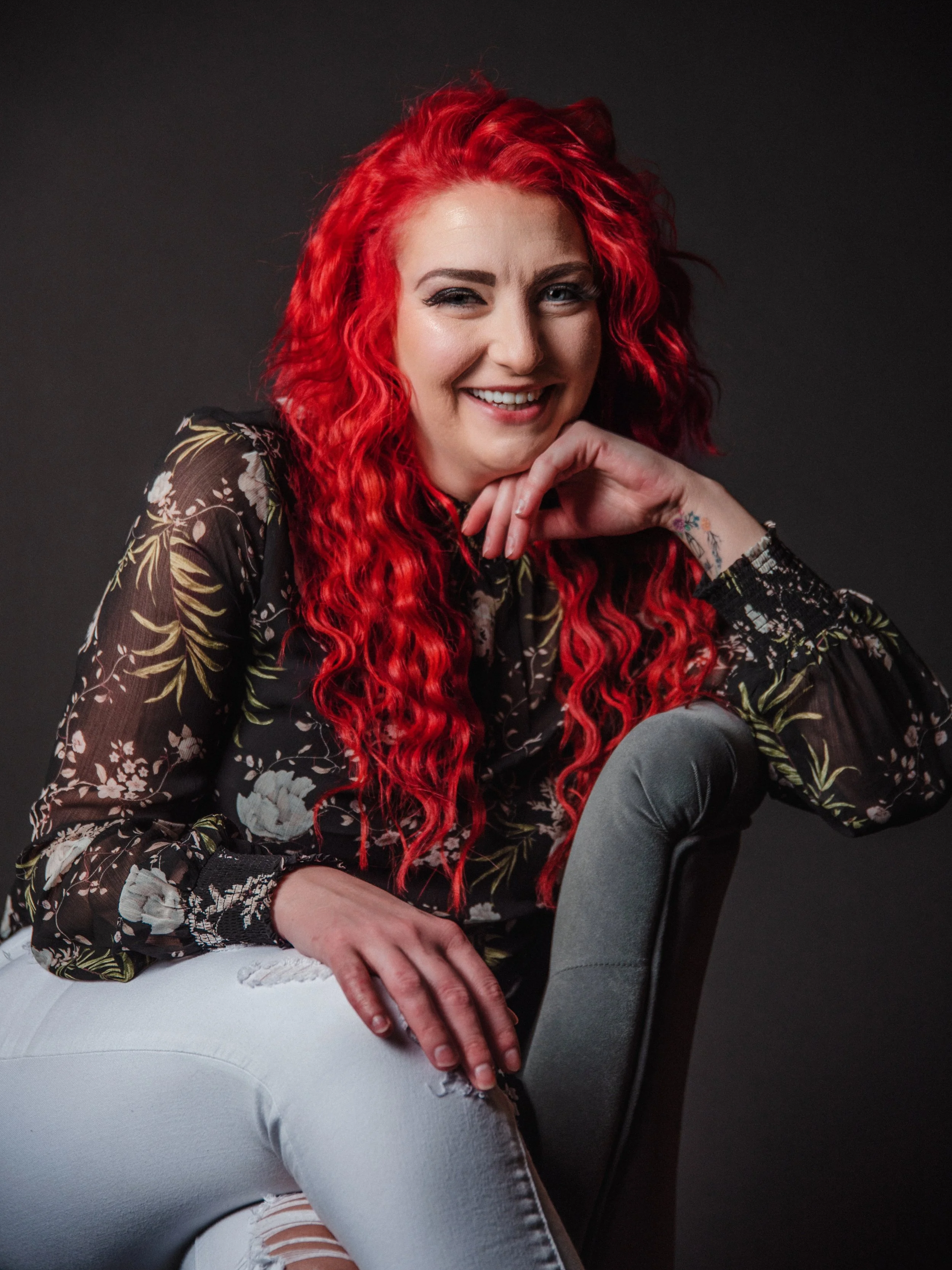 A woman with bright red, curly hair smiling and posing, sitting on a gray chair against a dark gray background, wearing a black floral blouse and distressed white jeans.