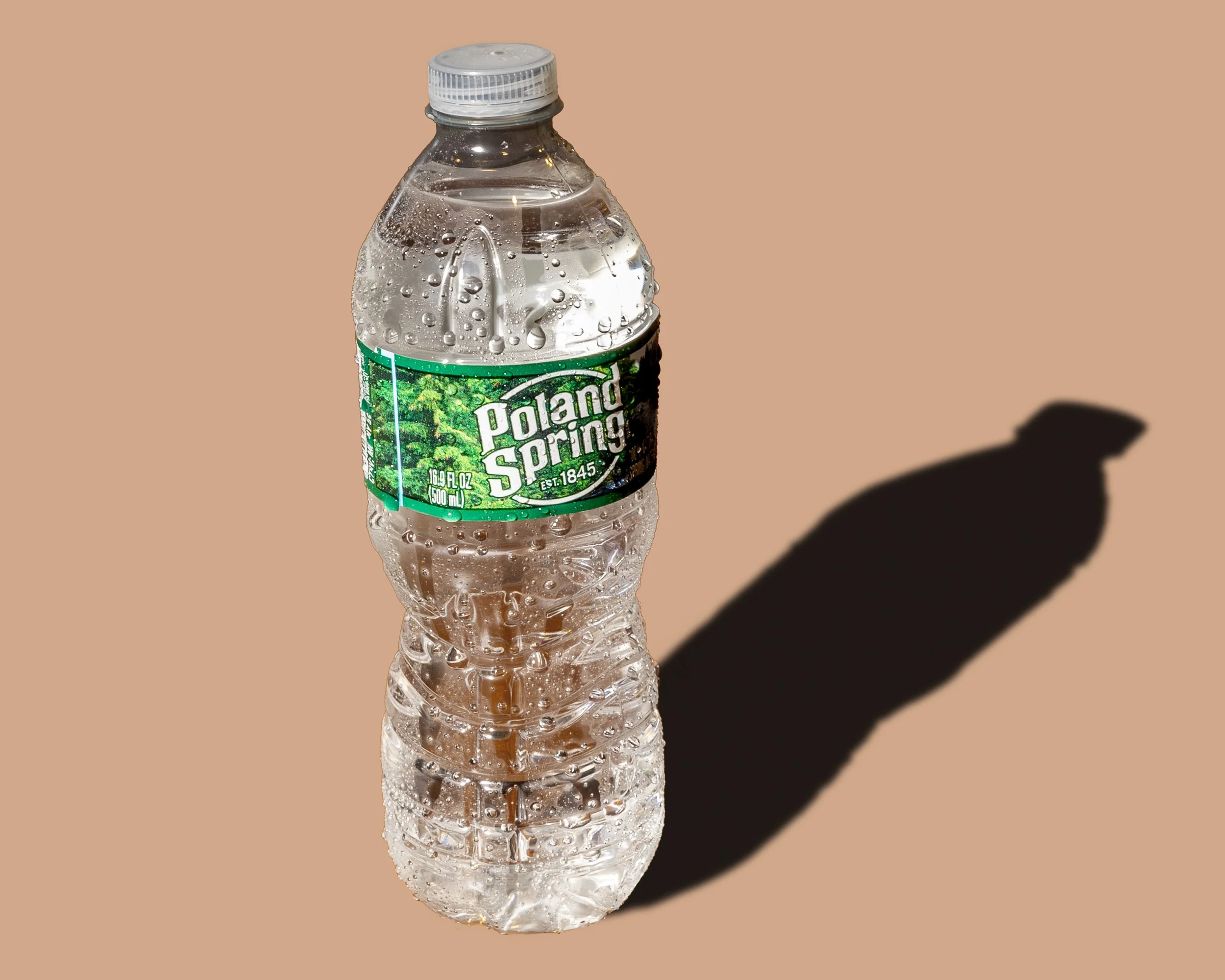 A clear plastic bottle of Poland Spring water with condensation on the outside. The bottle is placed on a surface, casting a shadow to the right, against a light brown background.
