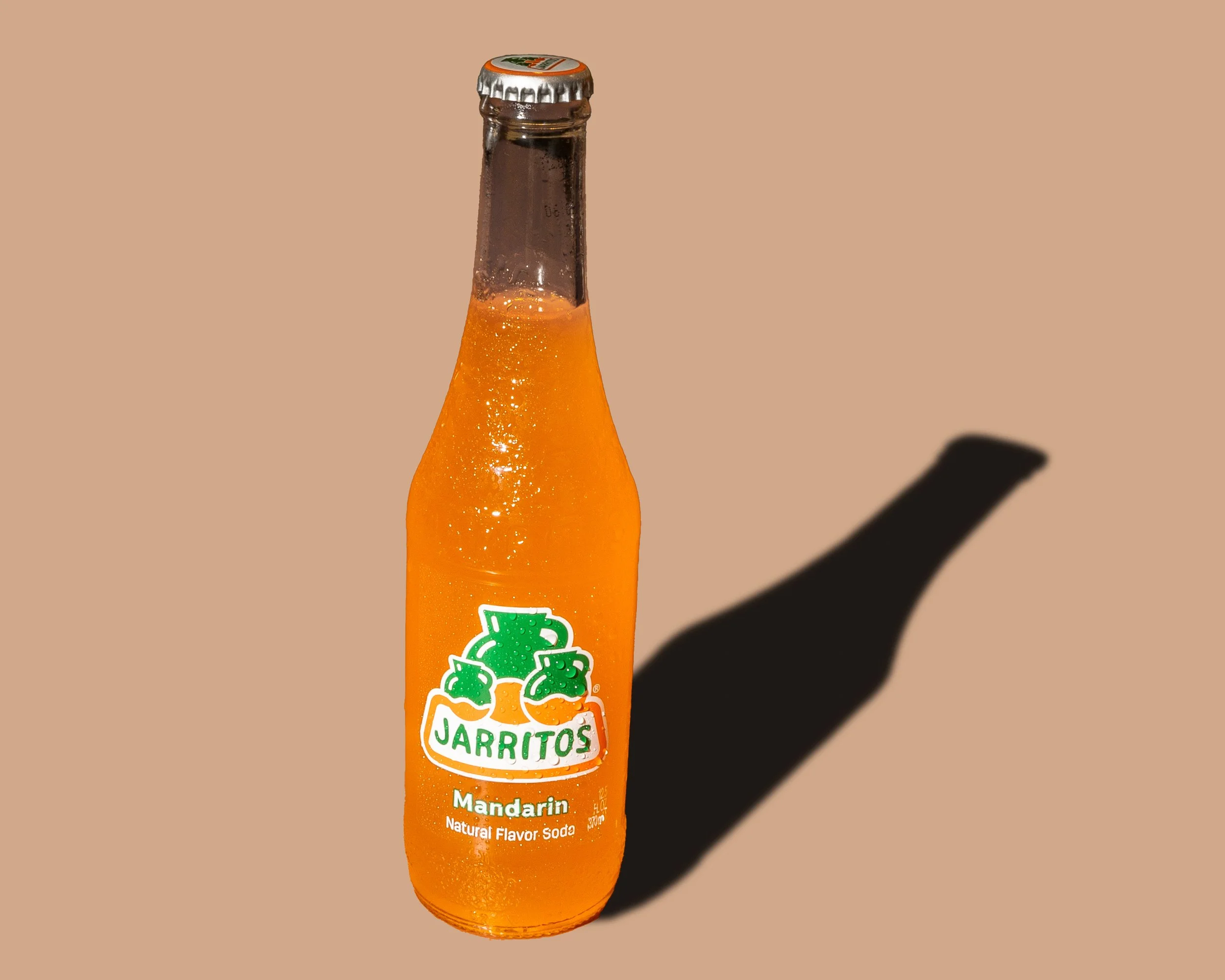 A glass bottle of Jarritos Mandarina soda with an orange-colored liquid inside, featuring a green and white logo on the label and a silver bottle cap. The bottle is casting a shadow on a plain beige background.