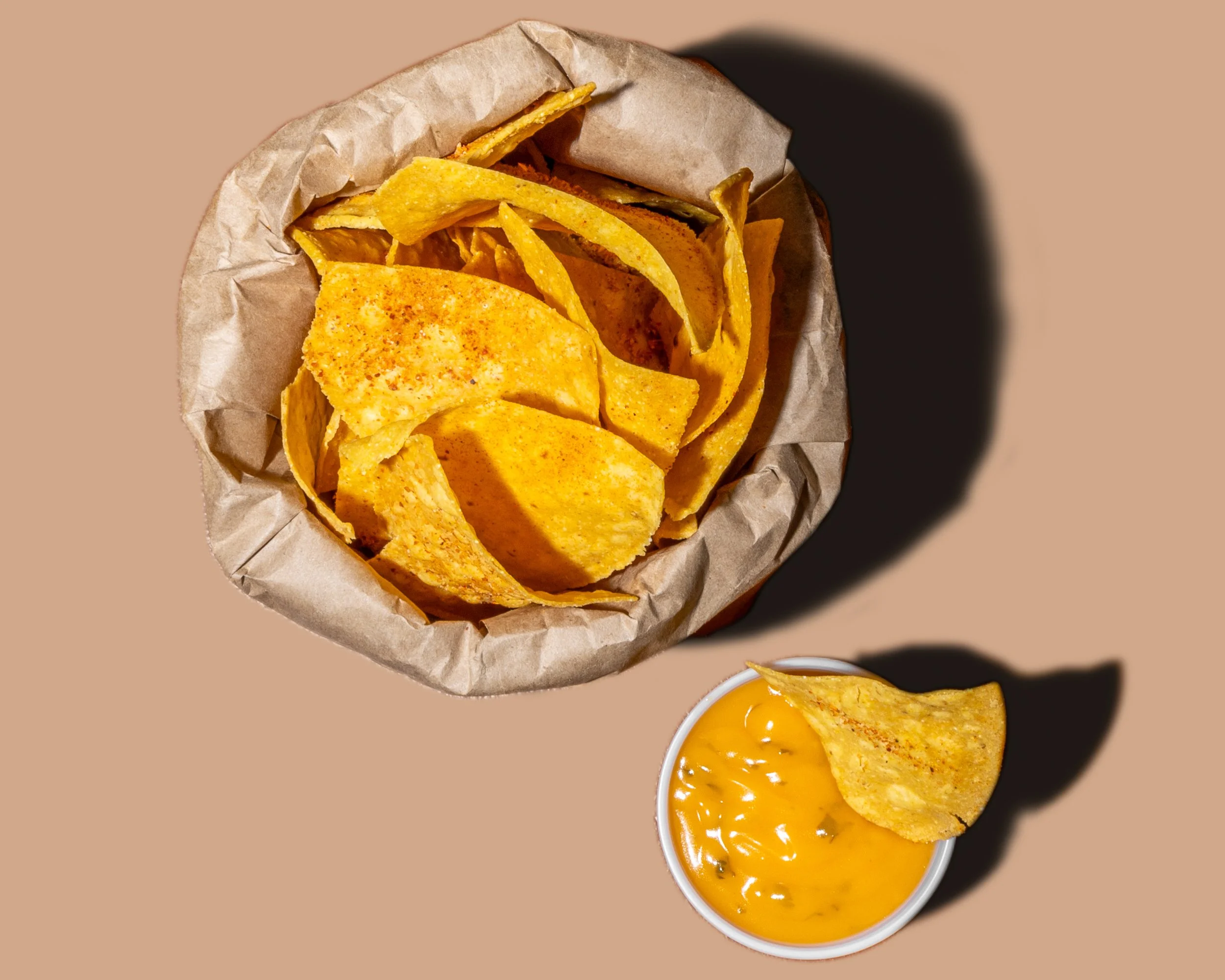 A paper wrapper containing potato chips, a small cup of nacho cheese sauce with a chip dipped in it, and a side of crispy tortilla chips on a beige background.
