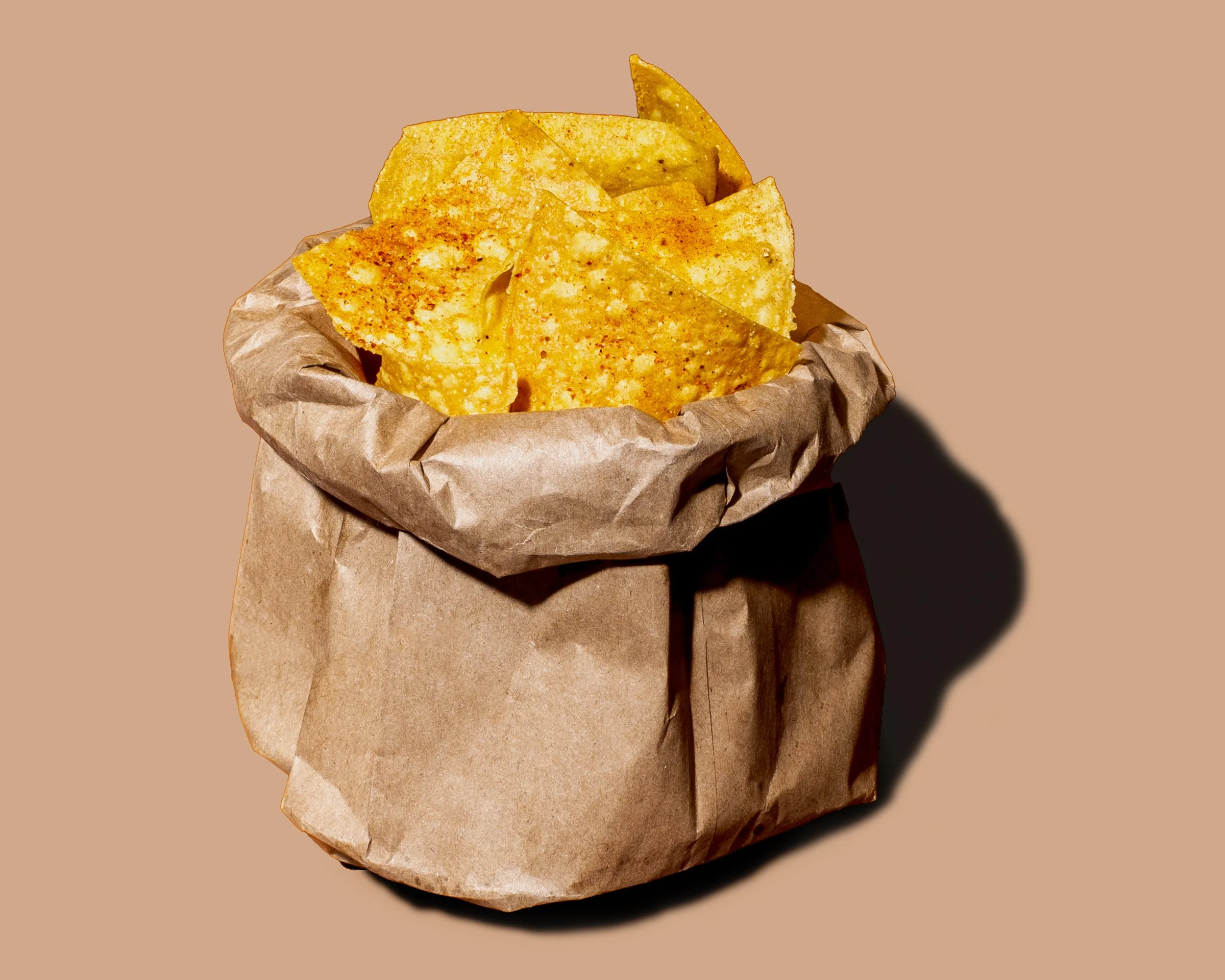 A brown paper bag filled with yellow tortilla chips against a beige background.