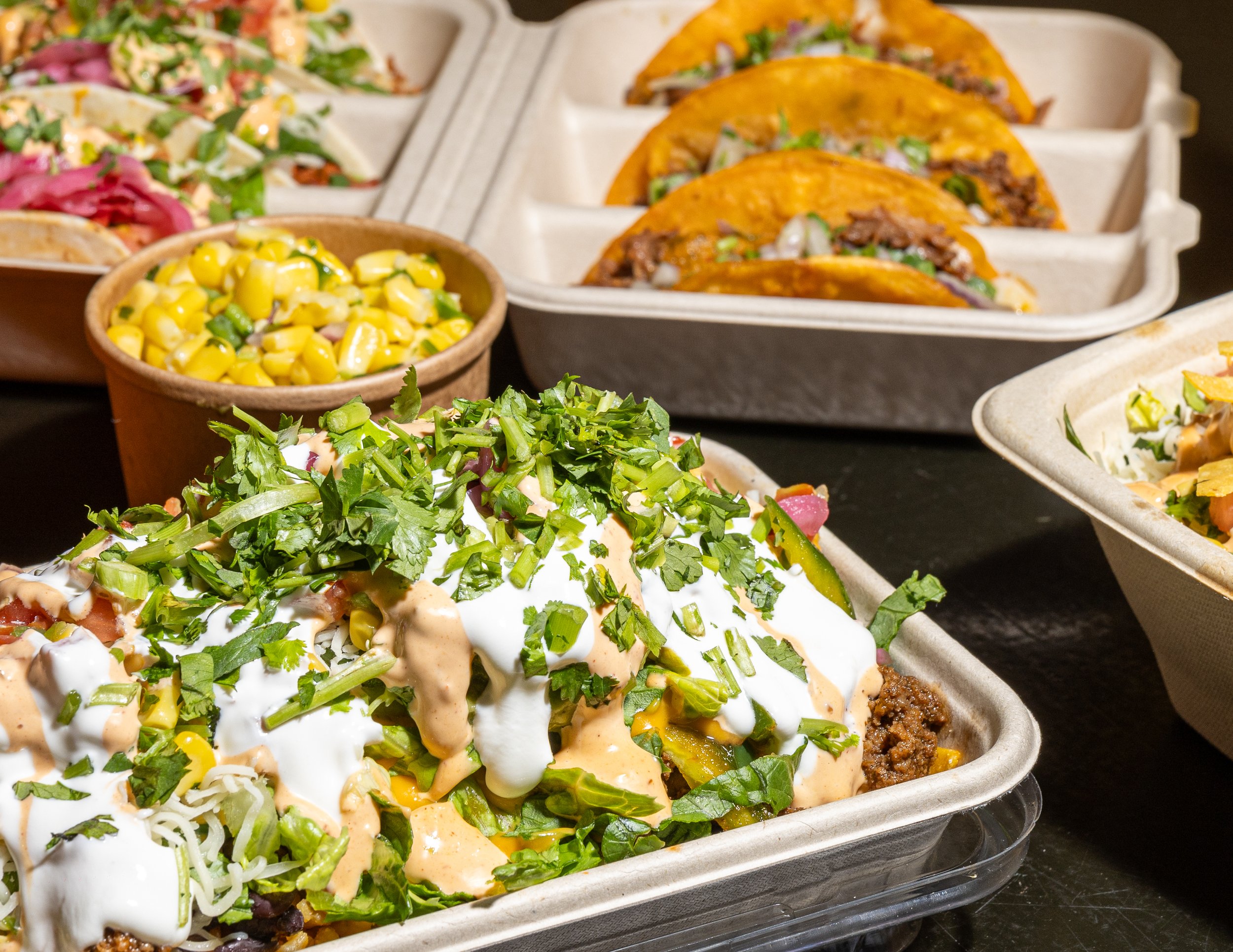 A tray of Mexican food featuring tacos, a bowl of corn salad, and salads topped with cilantro, cheese, and sauces.