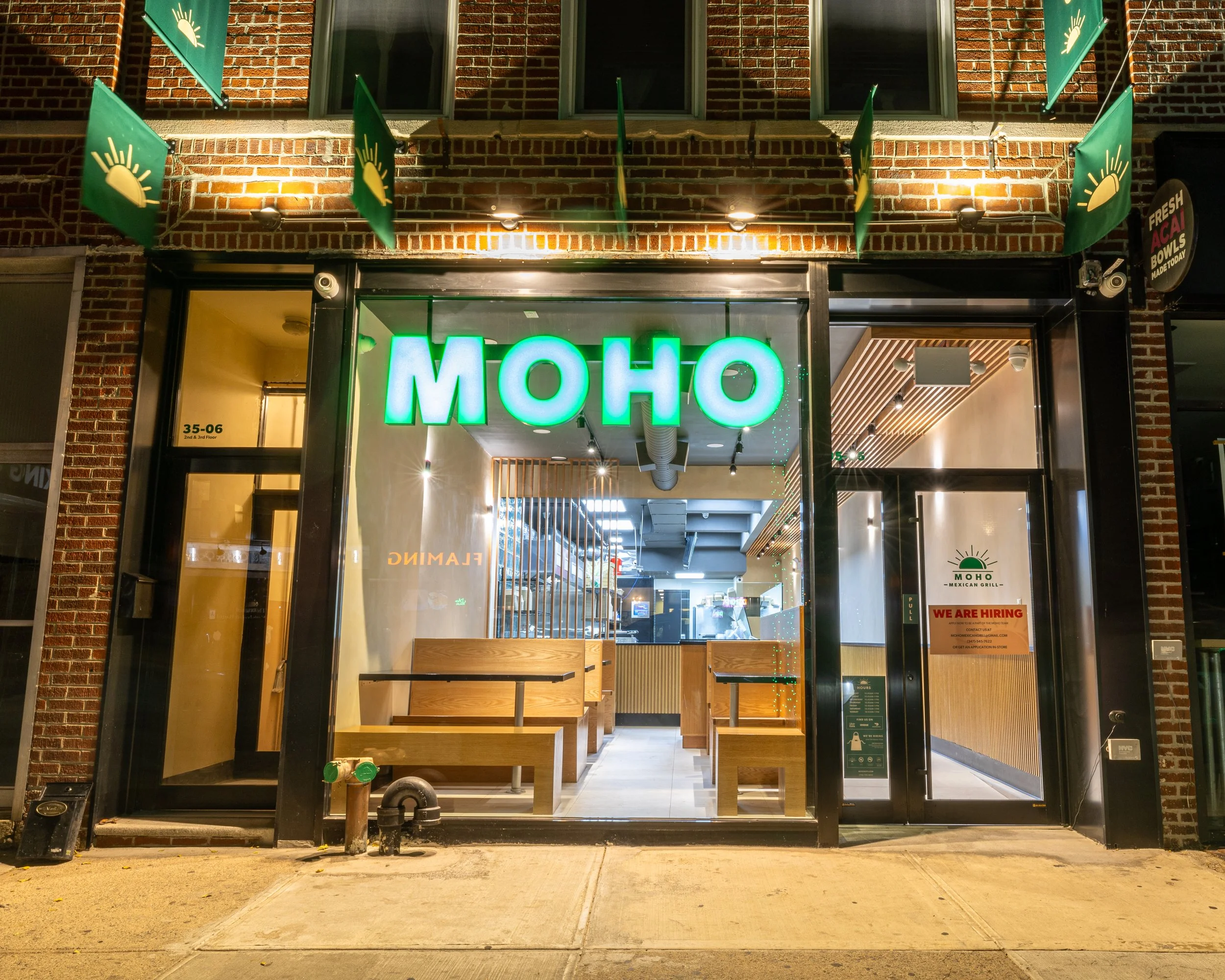 Night view of the storefront of MOHO Mexican Grill with illuminated green sign and green flags with a yellow sun logo hanging outside. Interior visible through glass doors and windows, with wooden tables and ceiling decor. A 'We Are Hiring' sign is on the right side.