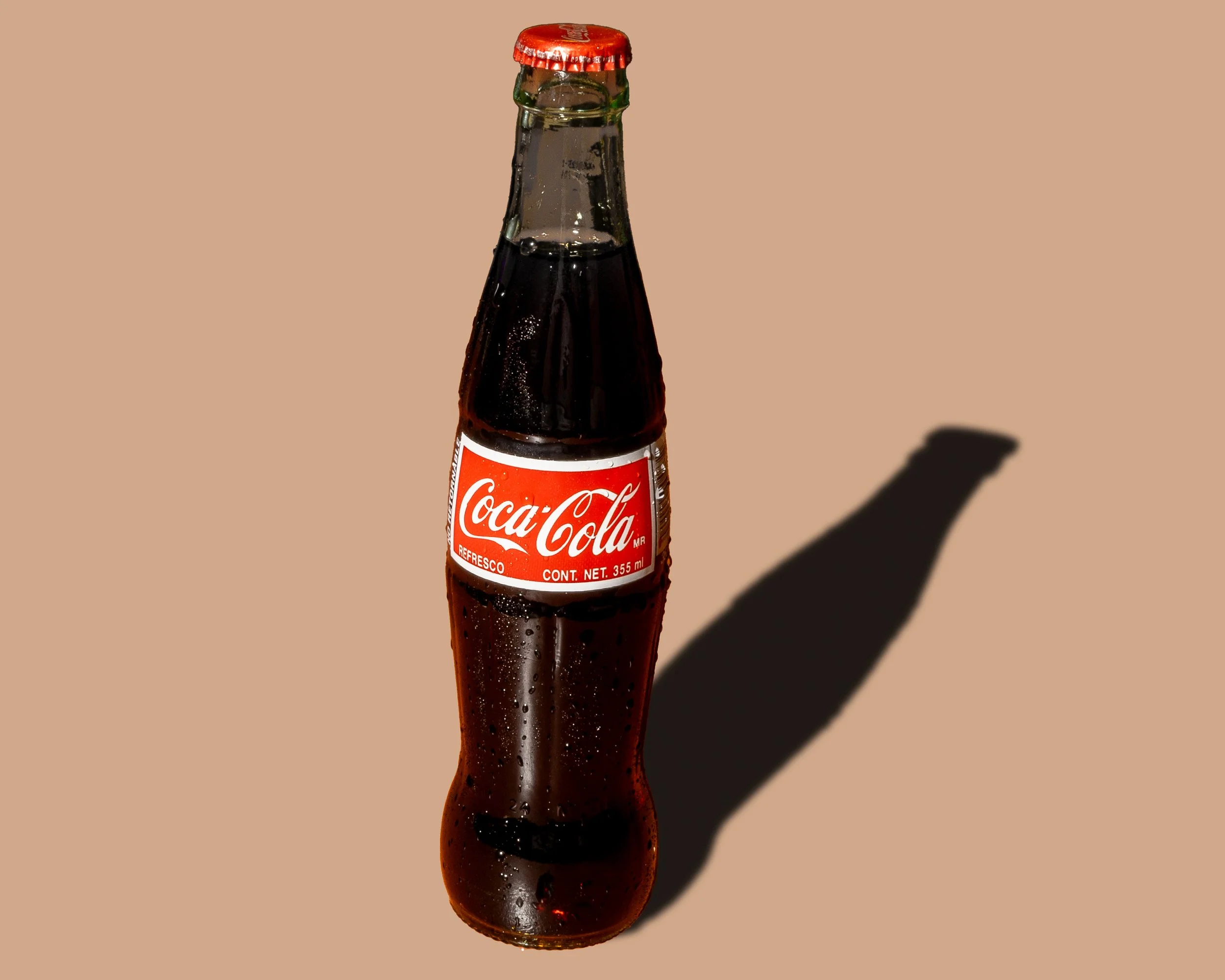 A glass Coca-Cola bottle with condensation, labeled as 355 ml, casting a shadow on a beige background.