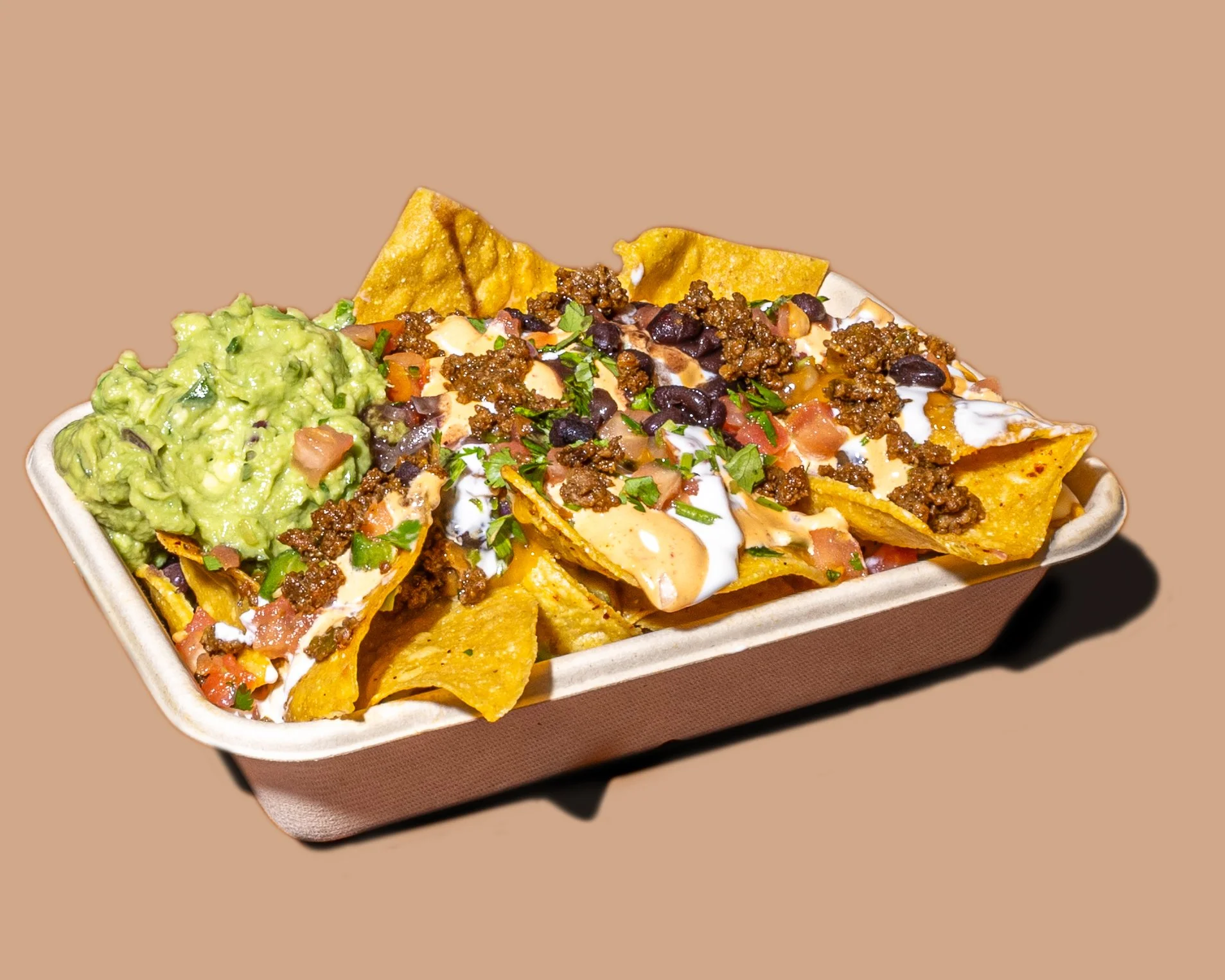 Close-up of a tray of nachos topped with ground beef, black beans, shredded cheese, sour cream, guacamole, diced tomatoes, chopped cilantro, and crushed tortilla chips on a brown background.