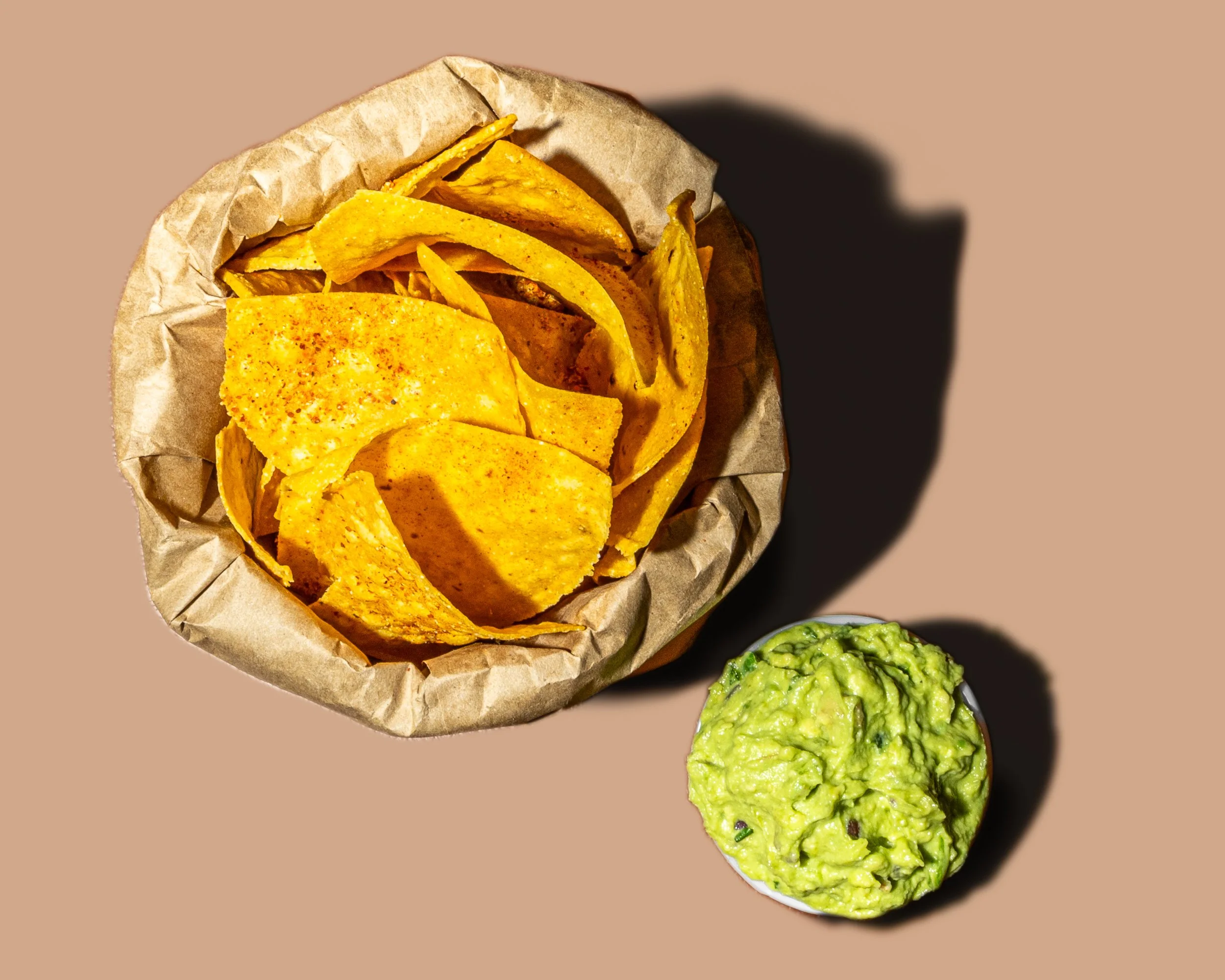 A paper wrapper filled with yellow corn tortilla chips, accompanied by a small bowl of guacamole on a neutral background.