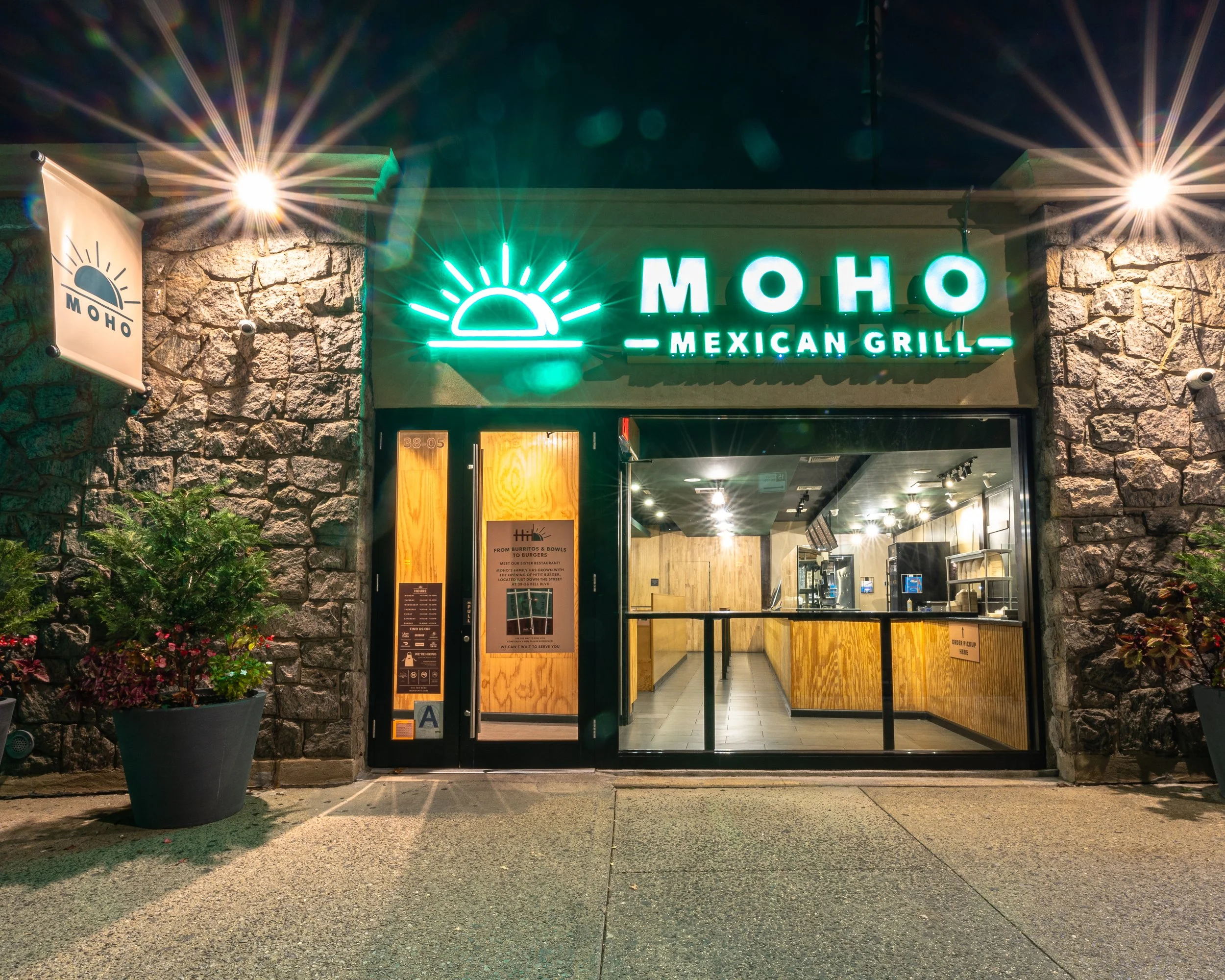 Night view of the entrance to Moho Mexican Grill, featuring a neon sign with a sun and horizon logo, stone facade, and glass door, with plants outside.