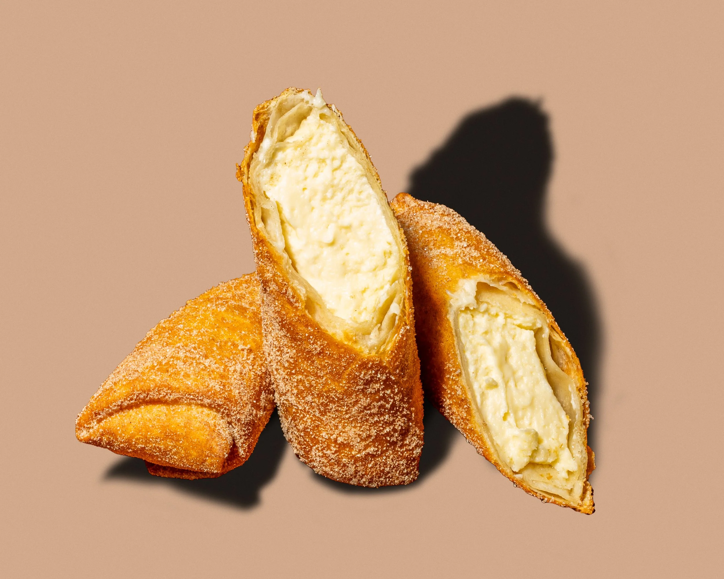 Two deep-fried cheese sticks with a crispy, golden-brown coating, one cut in half showing a melted cheese interior, placed on a light-colored background.