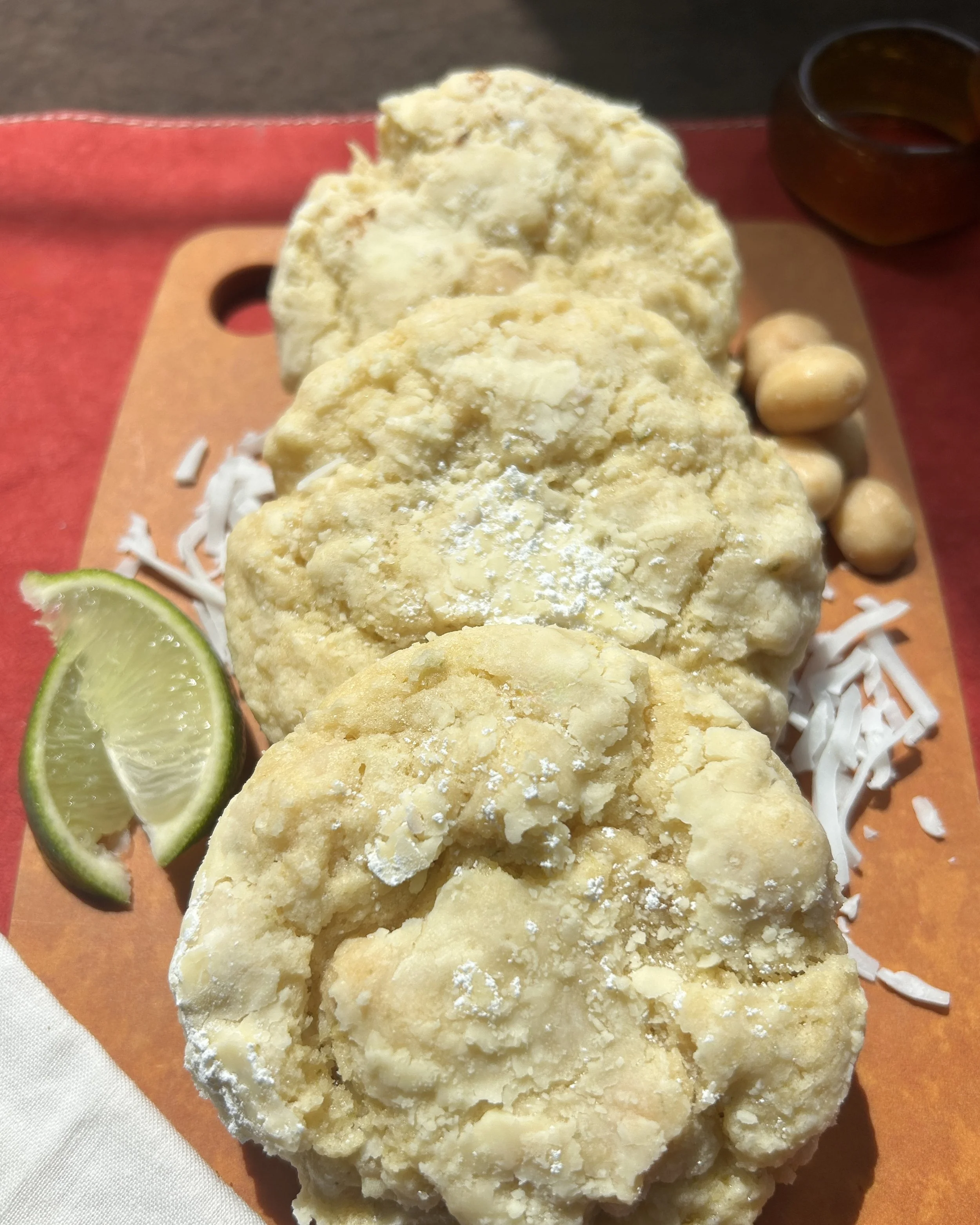 Close-up of three coconut lime macadamia nut cookies on a wooden platter, garnished with lime wedges and shredded white coconut.