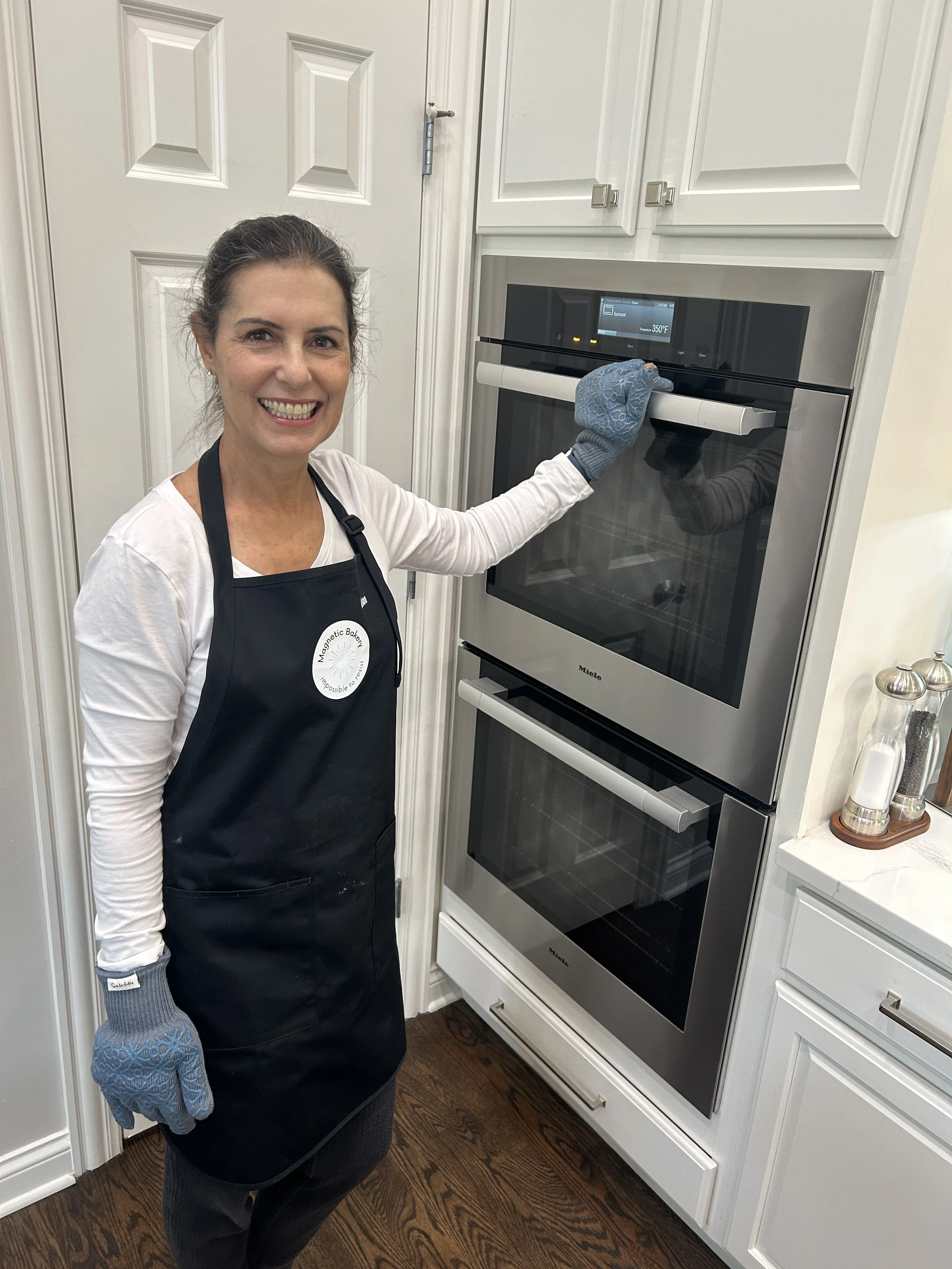 Alison, the founder wearing a black Magnetic Bakery apron and blue oven glove is standing in a kitchen, smiling, and using an oven.