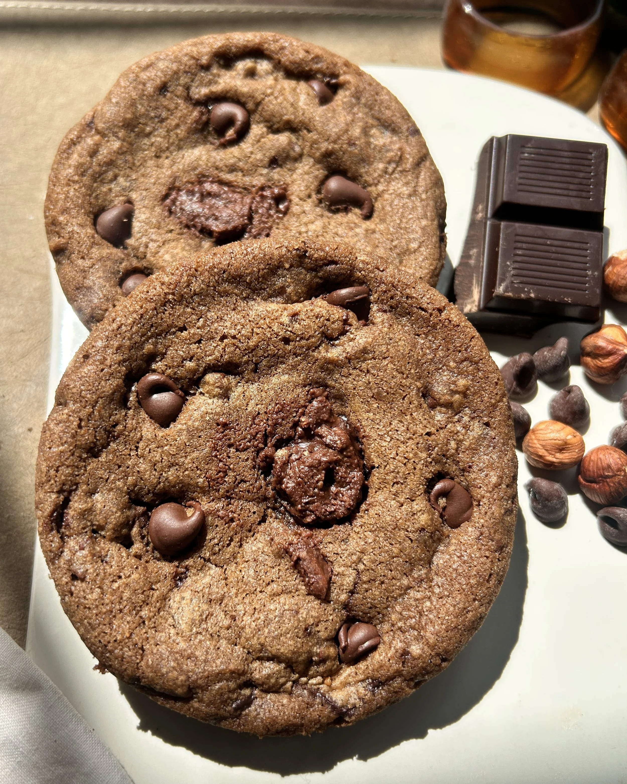 Close-up of two espresso chocolate hazelnut cookies on a white plate with scattered chocolate hazelnuts in the background.