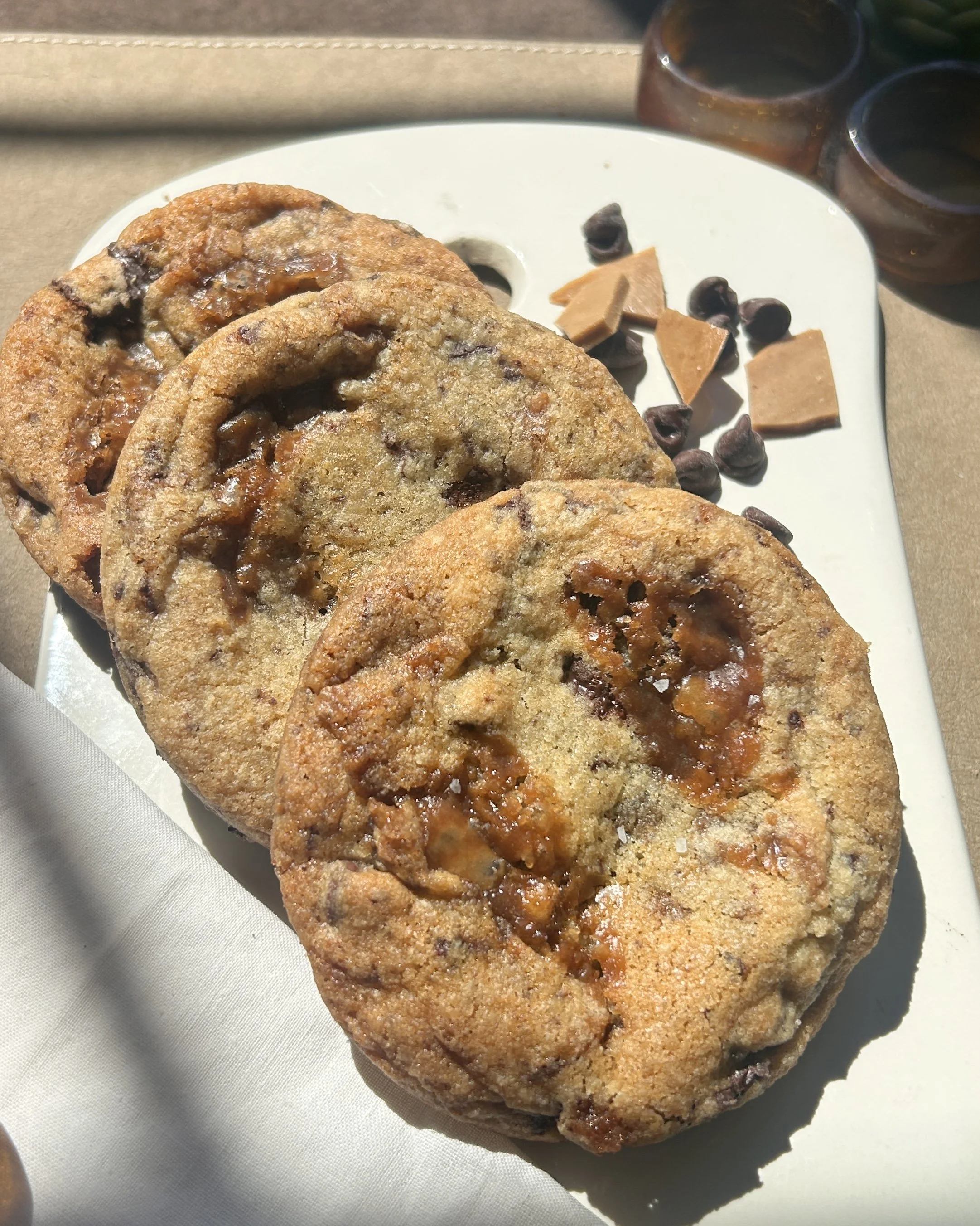 Close-up of three chocolate chip cookies with melting toffee inside, placed on a white oval dish. In the background, two brown glass napkin rings on a brown placemat.