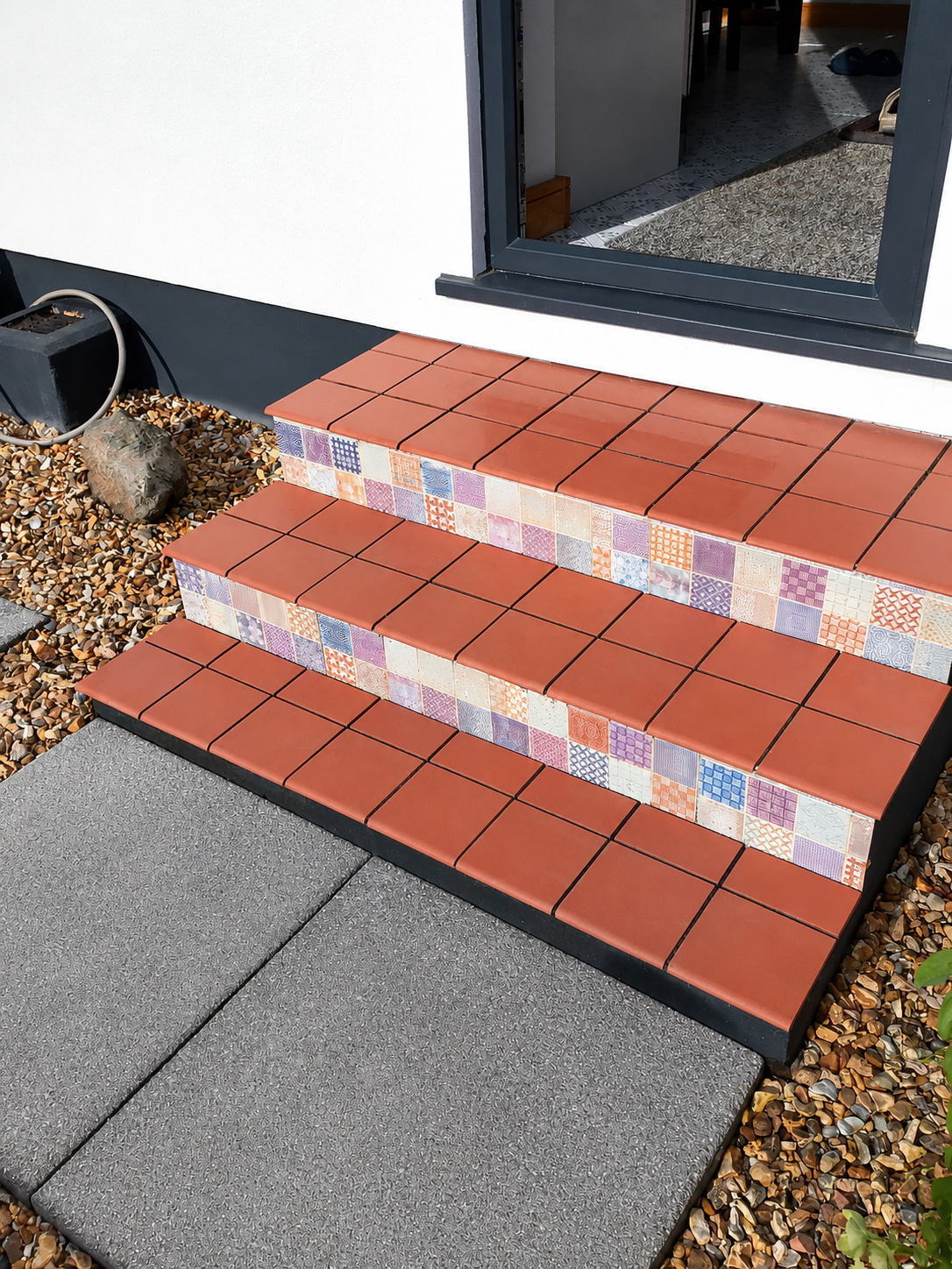 Decorative outdoor staircase with three steps, featuring red tiles on the top and sides with colorful patterned tiles on the front, next to a door and gravel landscaping.