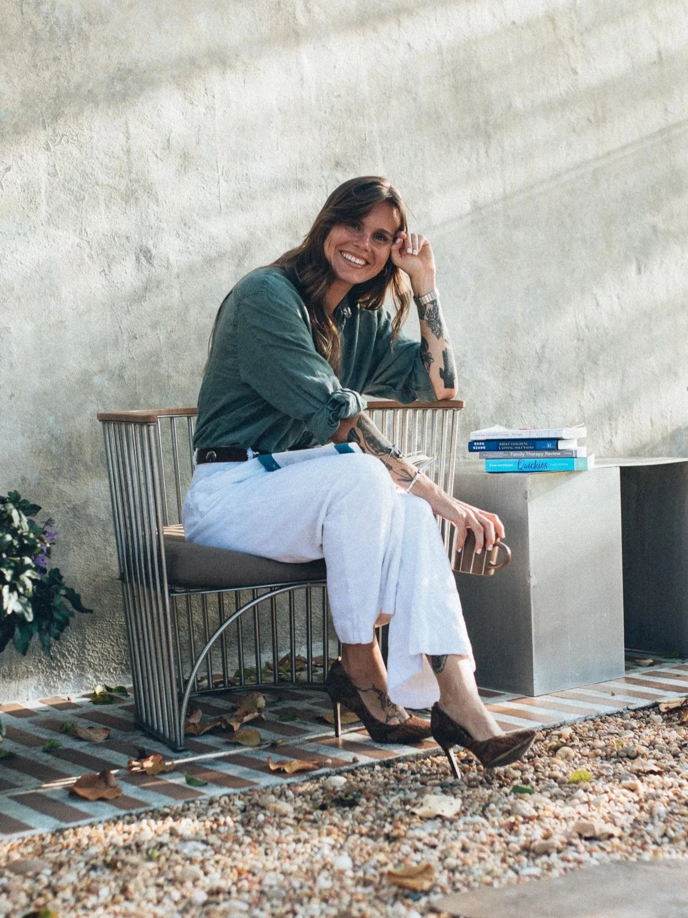 A woman sitting on a modern metal bench outdoors against a concrete wall, smiling, with books and wearing a green long-sleeve top, white pants, and high-heeled shoes.