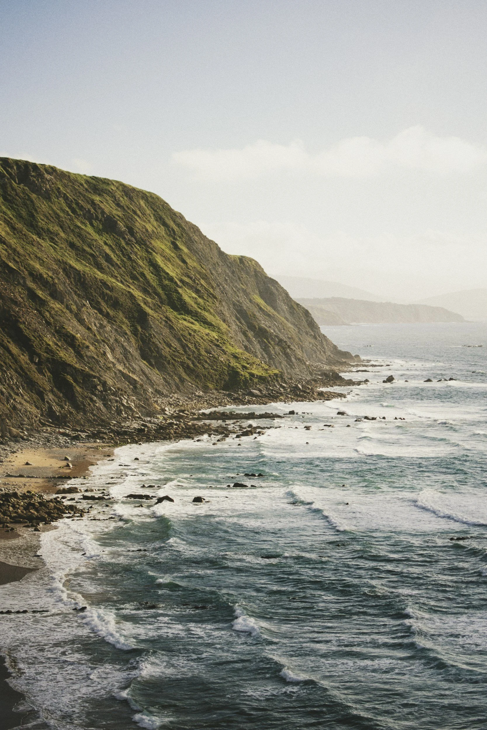 View of rugged green coastal cliffs with a sandy beach and ocean waves in the foreground, under a partly cloudy sky.